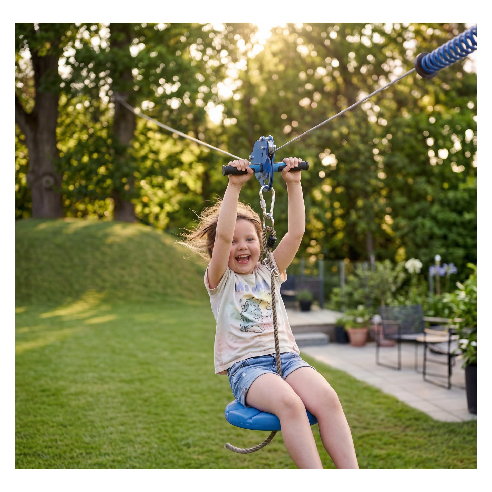 Das lächelnde Kind fährt die Kayoba-Seilbahn im Garten, trägt T-Shirt und Shorts. Bäume, Gras und Gartenmöbel im Sonnenlicht.