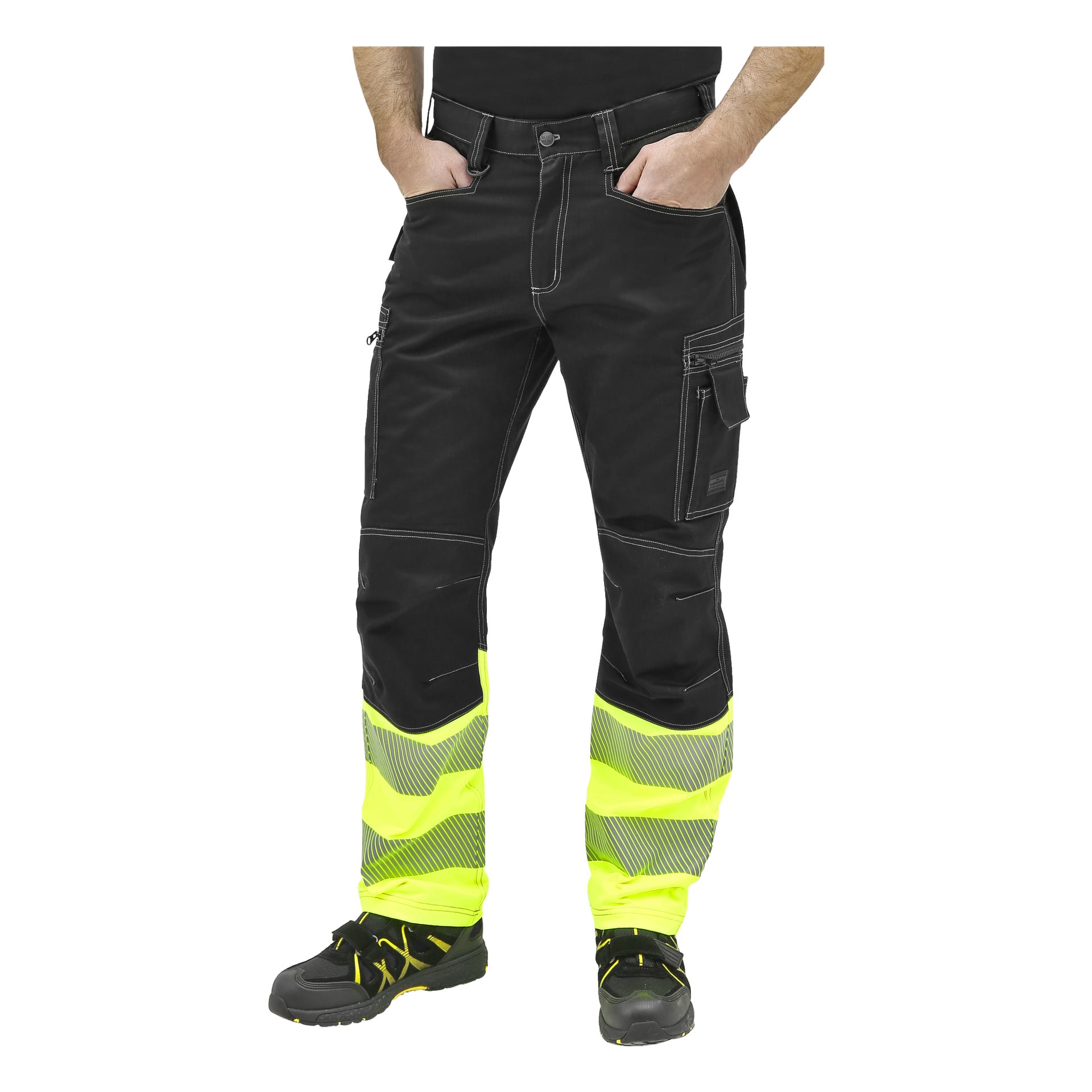 En person står med händerna i fickorna, bär Bluewear High Sight-byxor utan spikfickor som är svarta med gula high-sight och reflekterande silverband på underbenen, tillsammans med svarta arbetsskor med gula detaljer.