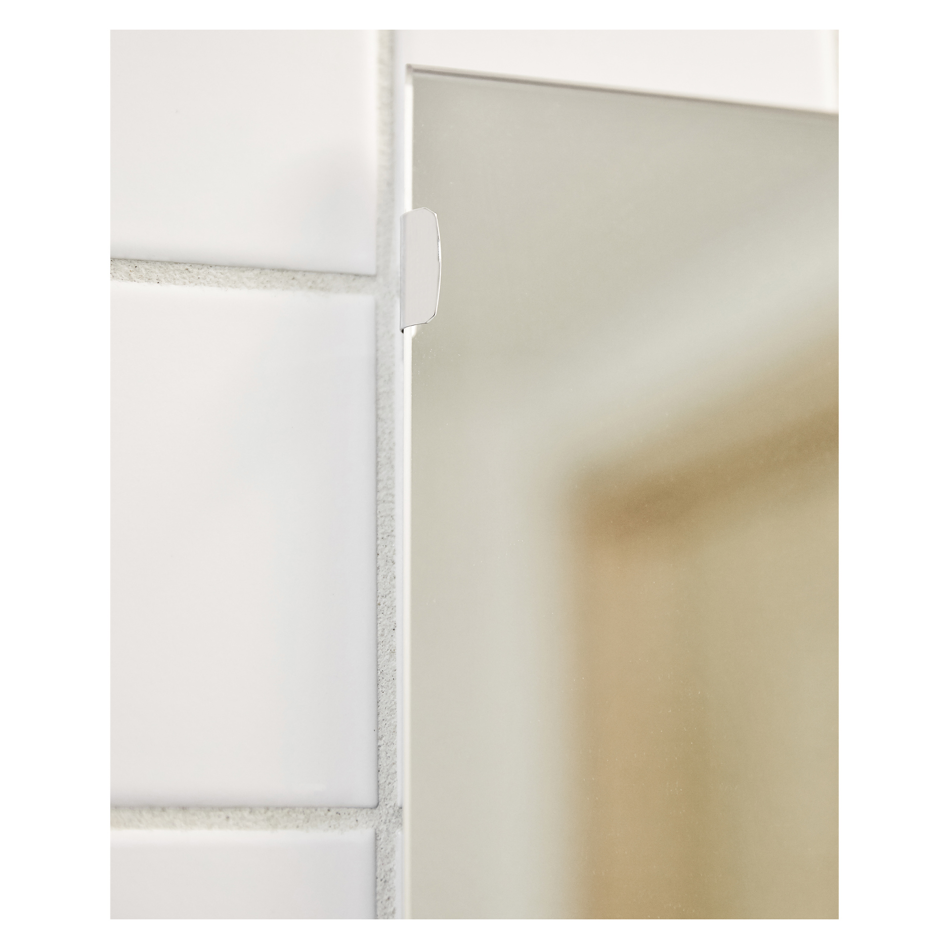 En detaljerad vy av kanten på en ramlös spegel fäst vid en vit kaklad vägg med en Hard Head Mirror-hållare, en del av en fyrdelad uppsättning. Höger sida av bilden visar en delvis suddig reflektion.