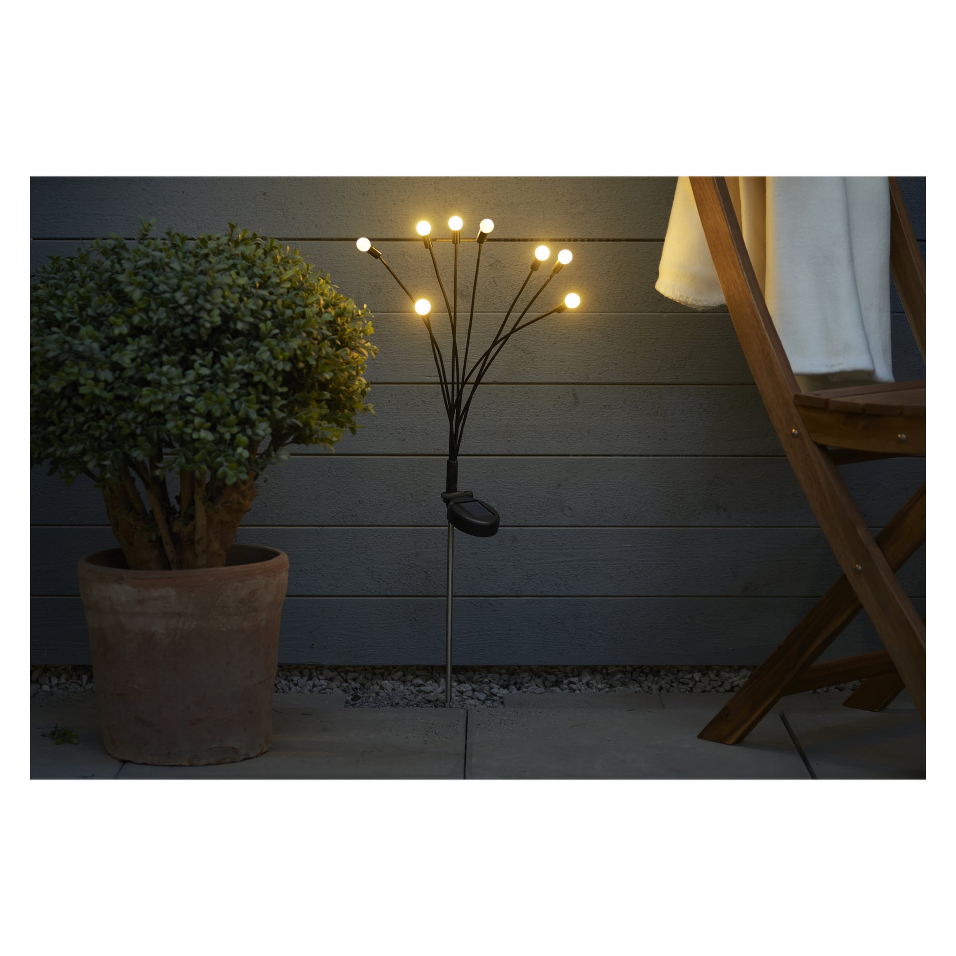 En krukväxt står bredvid Anslut solcellslampa LED Firefly, dess rad av lysande glödlampor lyser upp området bredvid en trästol på en uteplats med grå horisontell fasad bakom.
