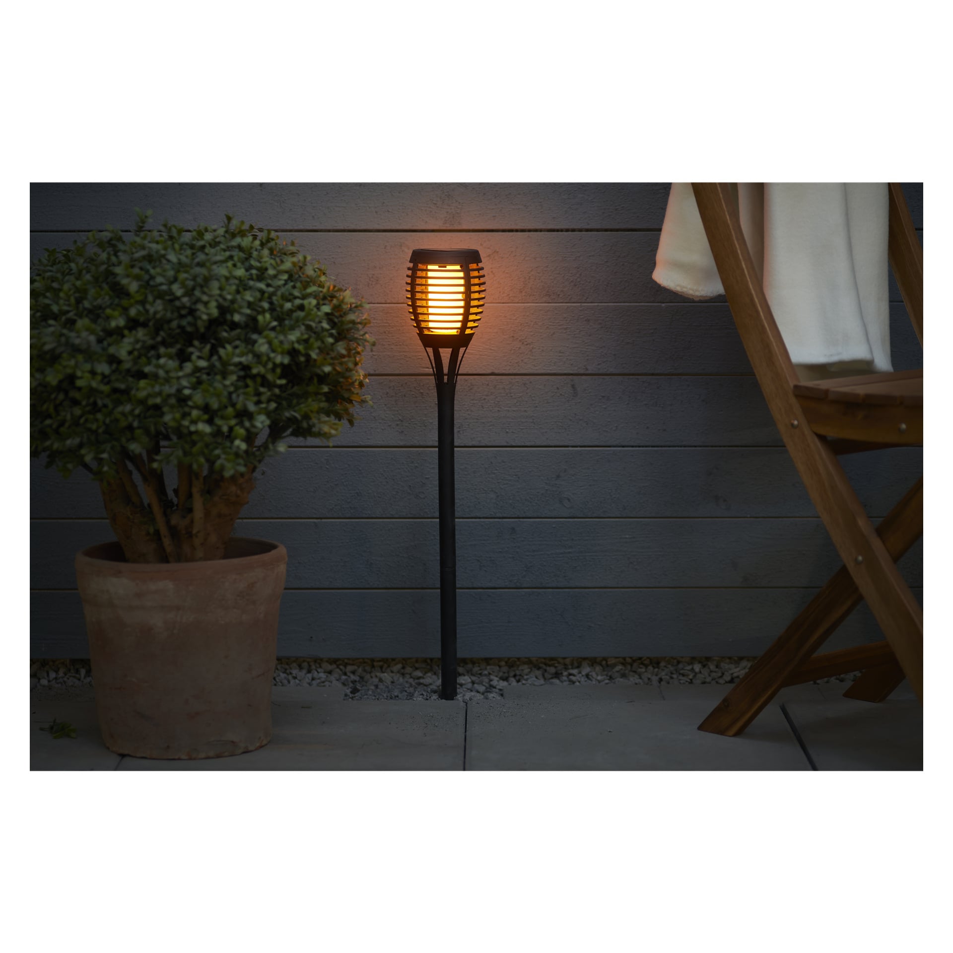 En krukväxt står på en uteplats bredvid en hög svart Anslut solcells-LED-lampa, som ger varm belysning framför ett grått trästaket. Till höger syns en del av en trästol med en vit duk draperad över.