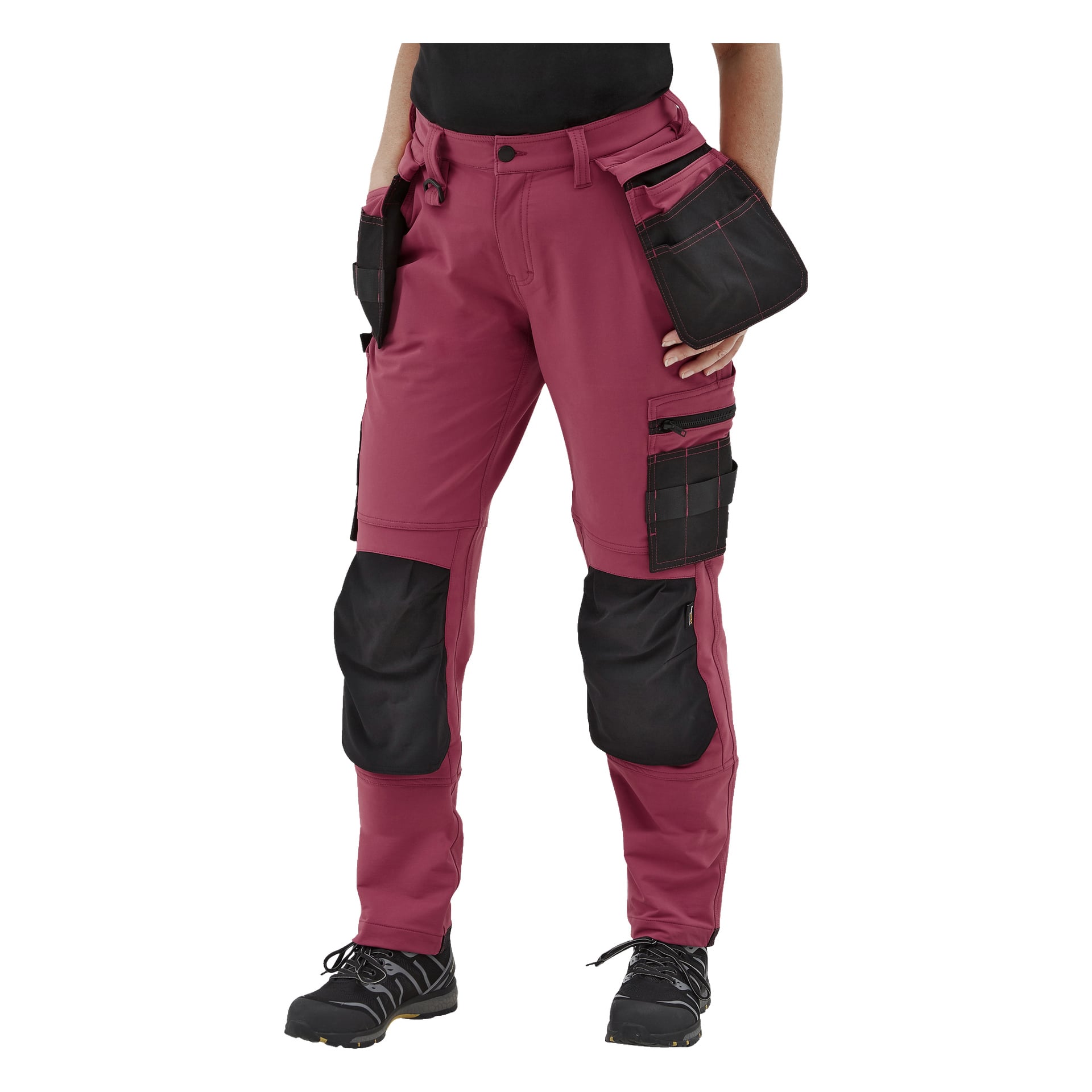 En person bär Bluewear Pro Workwear Women's work pants Stella i rött med svarta knäskydd, verktygsfickor och flera fickor, tillsammans med en svart skjorta och svarta arbetsskor.