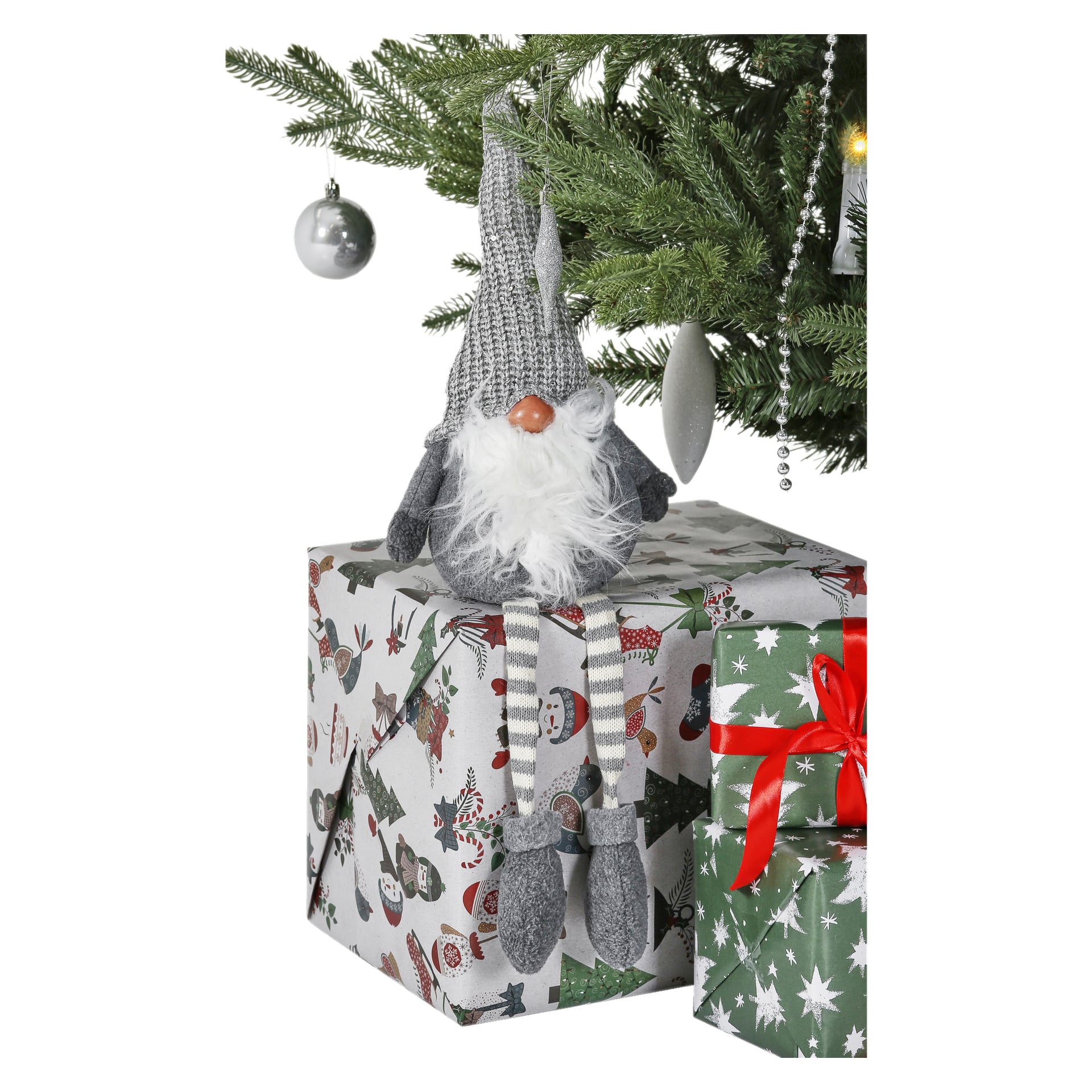 En tomtefigur med hängande ben från EKVIP sitter på en inslagen julklapp under en dekorerad gran, och det finns en annan present i grönt och rött i närheten.