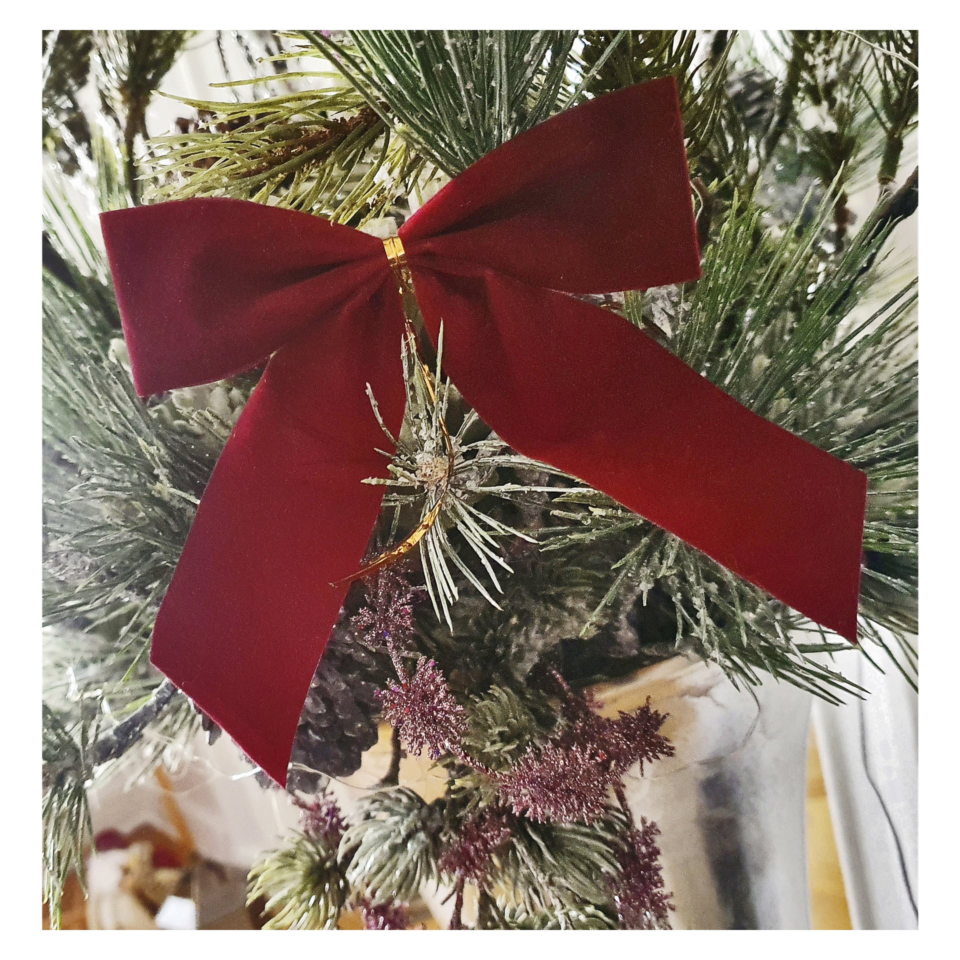 En närbild av en EKVIP Rosettes fyrpack röd sammetsrosett fäst vid konstgjorda gröna julgransgrenar, med kottar och olika prydnader bakom.