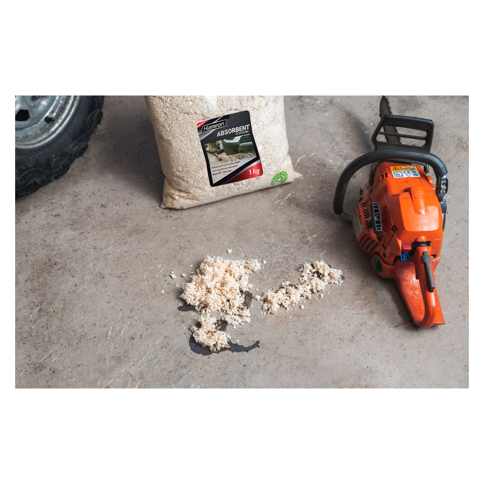 Hamron Absorbant från Biosorbe är staplad över ett spill på ett betonggolv. Bredvid den finns en påse märkt "Absorbent", en orange motorsåg, och en del av ett fordonsdäck dyker upp vid scenens kant.