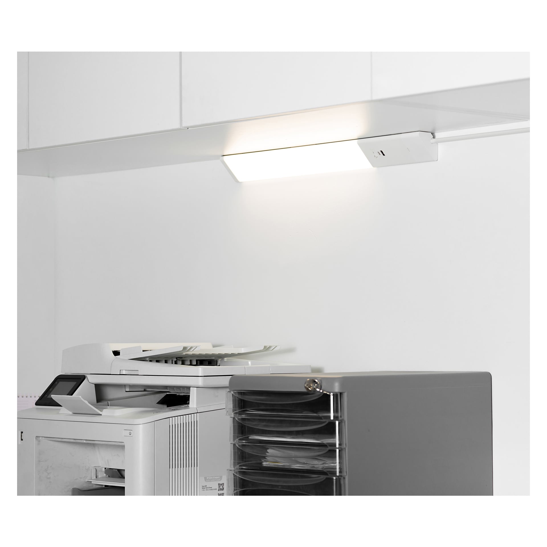 En vit multifunktionsskrivare står bredvid en grå organisatör med hyllor. Ovanför är Anslut Worktop-lampan installerad under ett skåp.