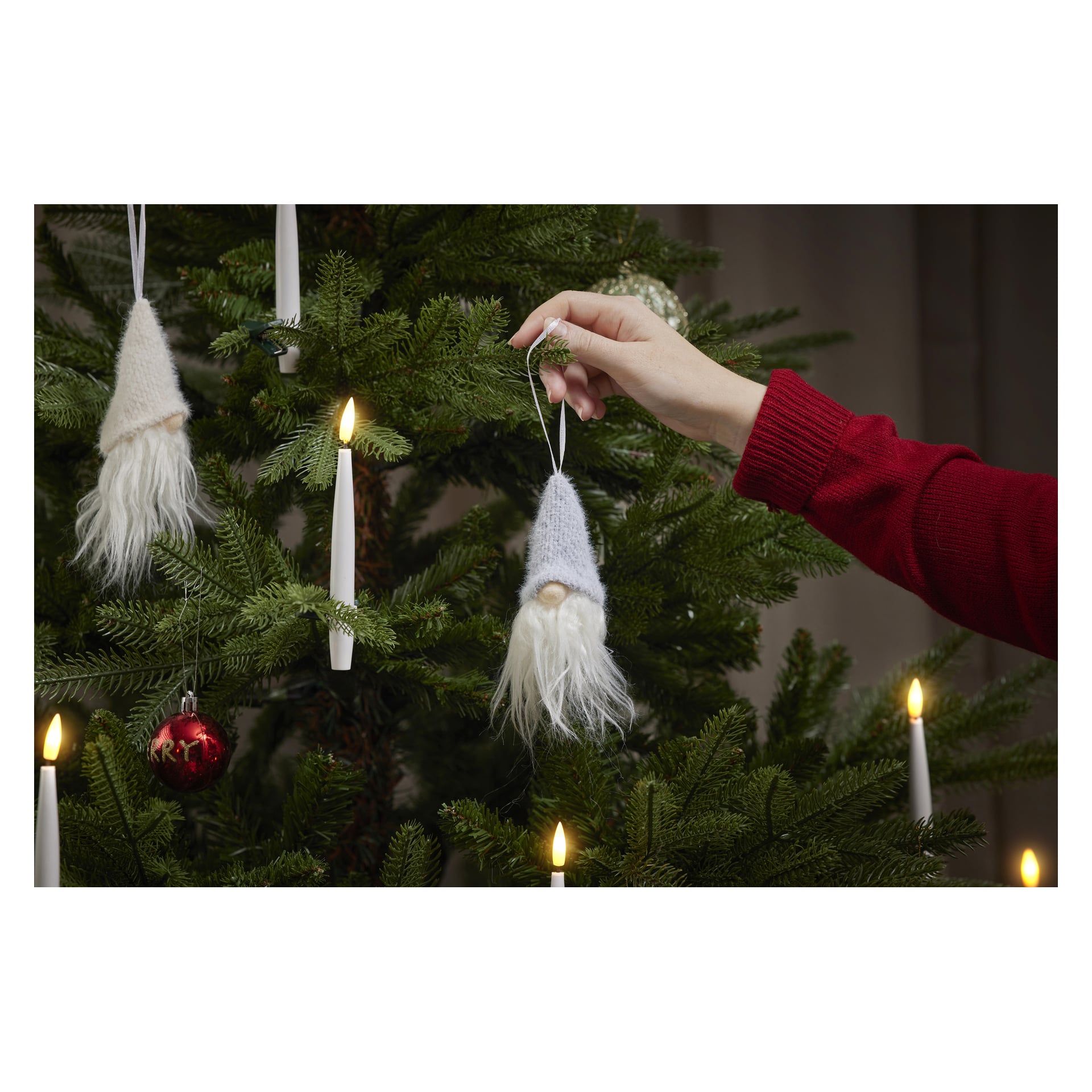 En person med röd ärm hänger en EKVIP-jultomte-prydnad med vitt skägg på en julgran dekorerad med ljus och kulor.