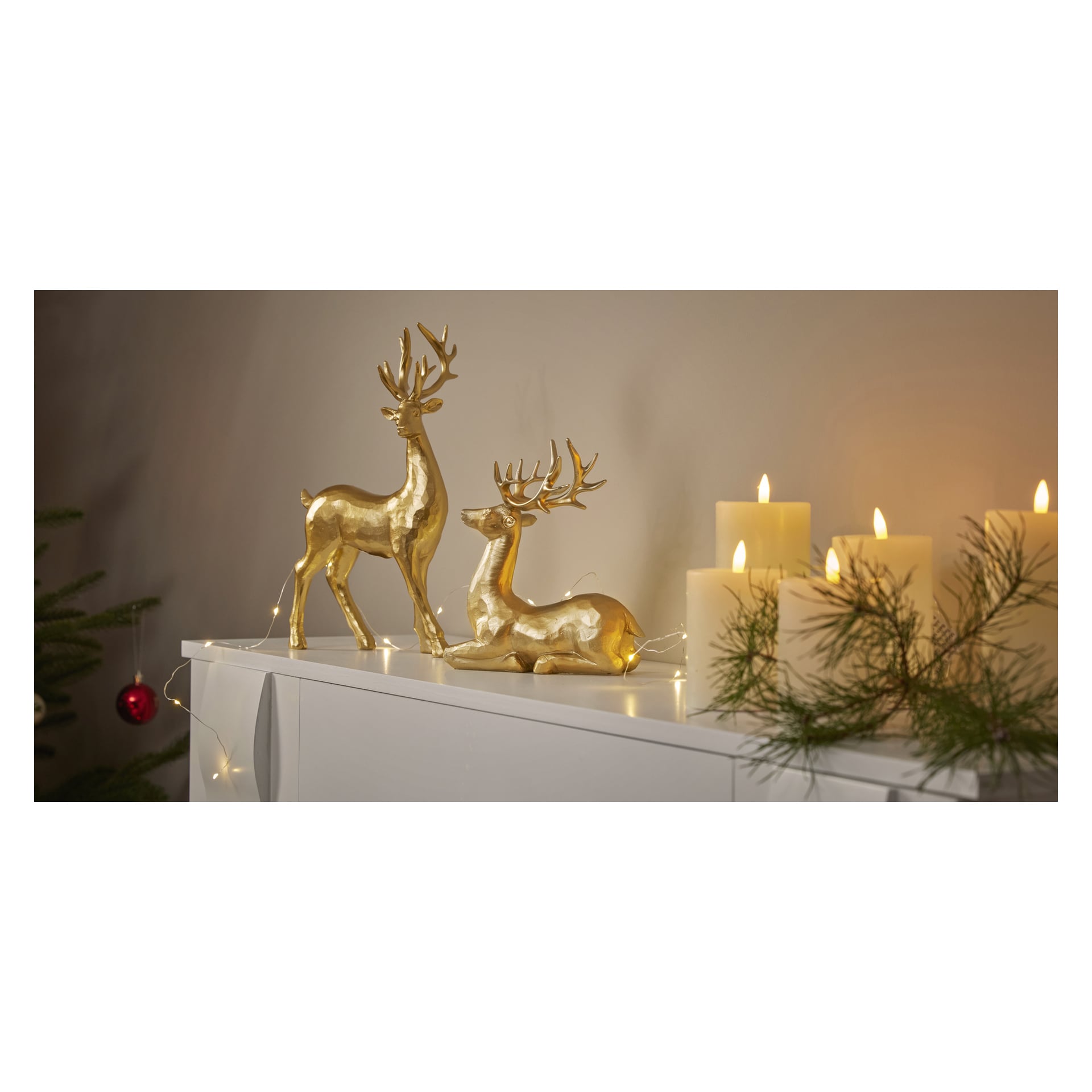 Två EKVIP Lying Deer Gold-figurer placeras på ett skåp, ackompanjerade av lysande pelarljus och tallgrenar. I bakgrunden finns ett träd med en hängande röd prydnad, vilket bidrar till den mysiga atmosfären.