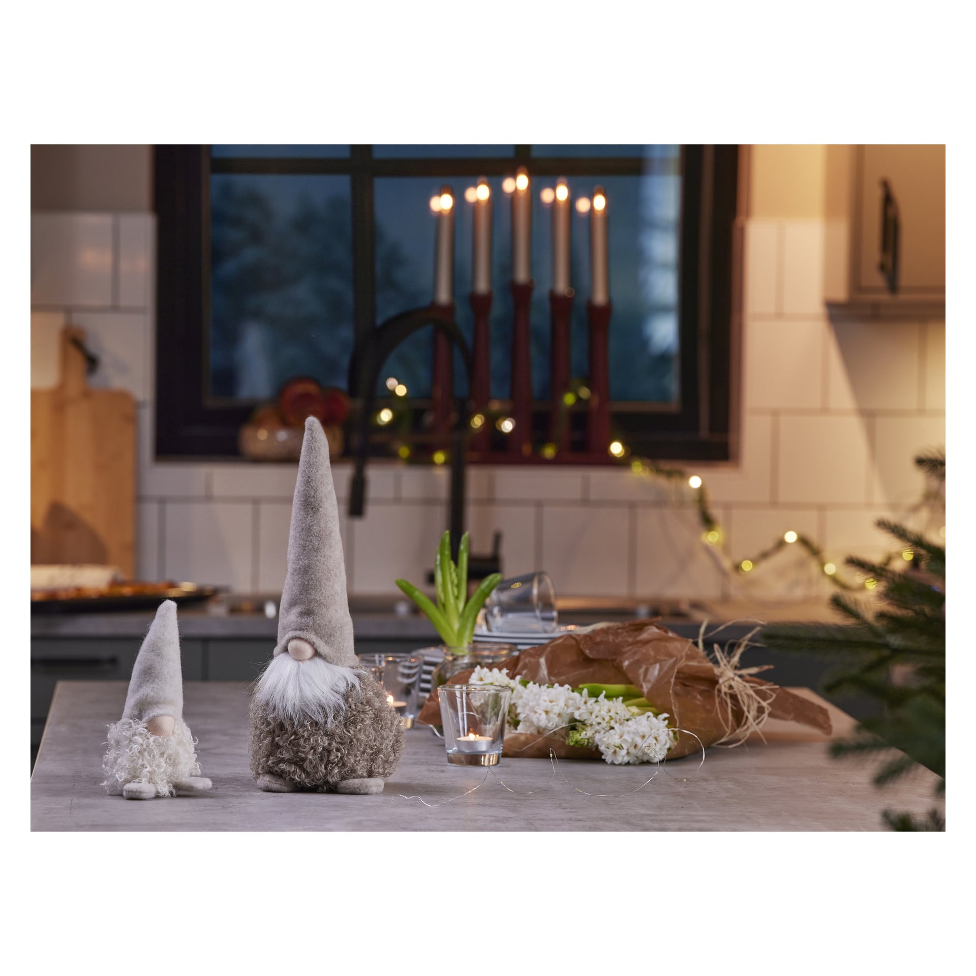 Två EKVIP tomtefigurer står på en köksbänk tillsammans med vita blommor, ett ljus och presentpapper. Bakom dem avslöjar ett fönster en ljusstake med sju brinnande ljus. Stämningen är varm och festlig.