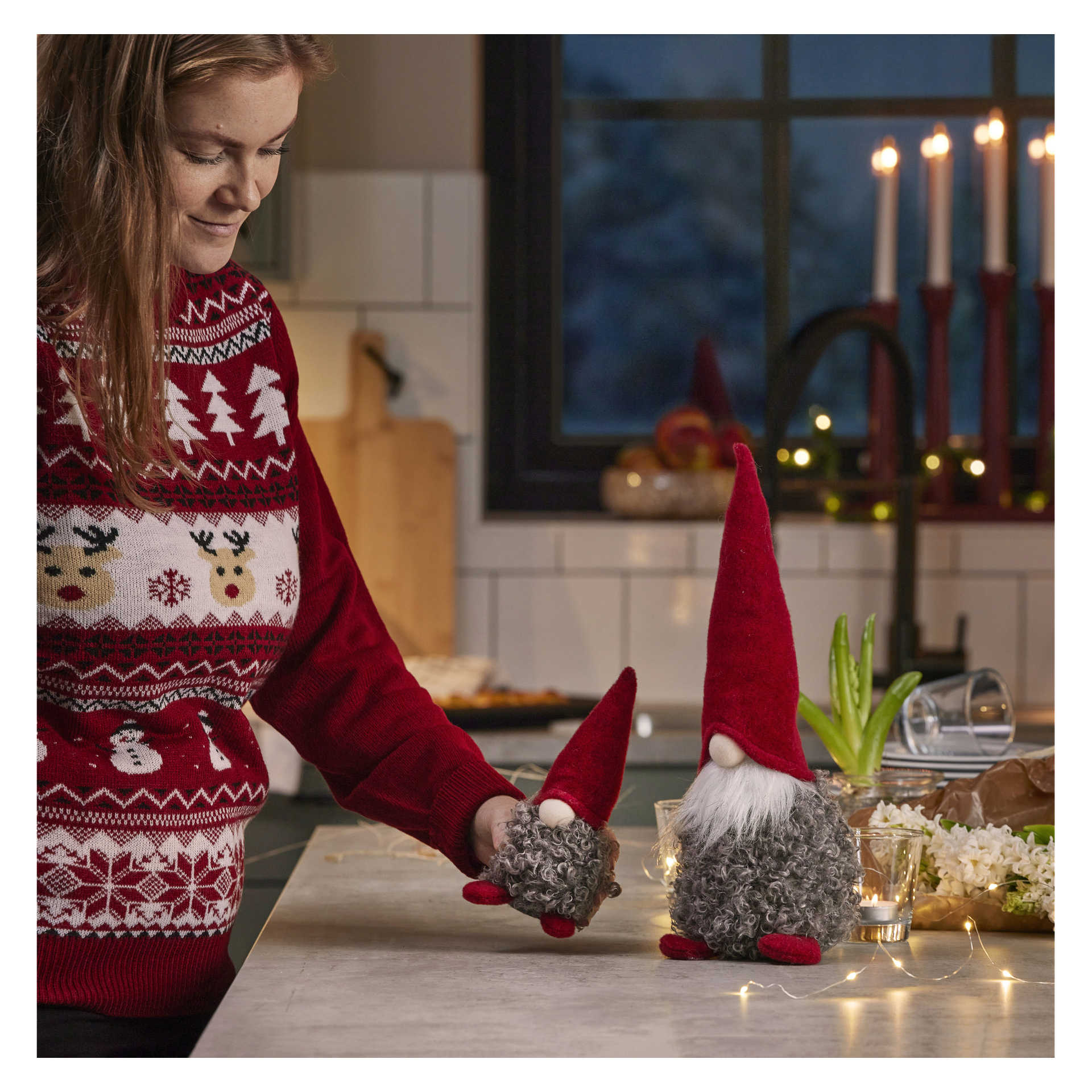En person med en röd jultröja står vid en köksbänk och håller i en liten EKVIP-tomtefigur. Disken visar också en större EKVIP-tomte, ljus, en krukväxt och ljusslingor. Fler tända ljus syns på fönsterbrädan bakom dem.