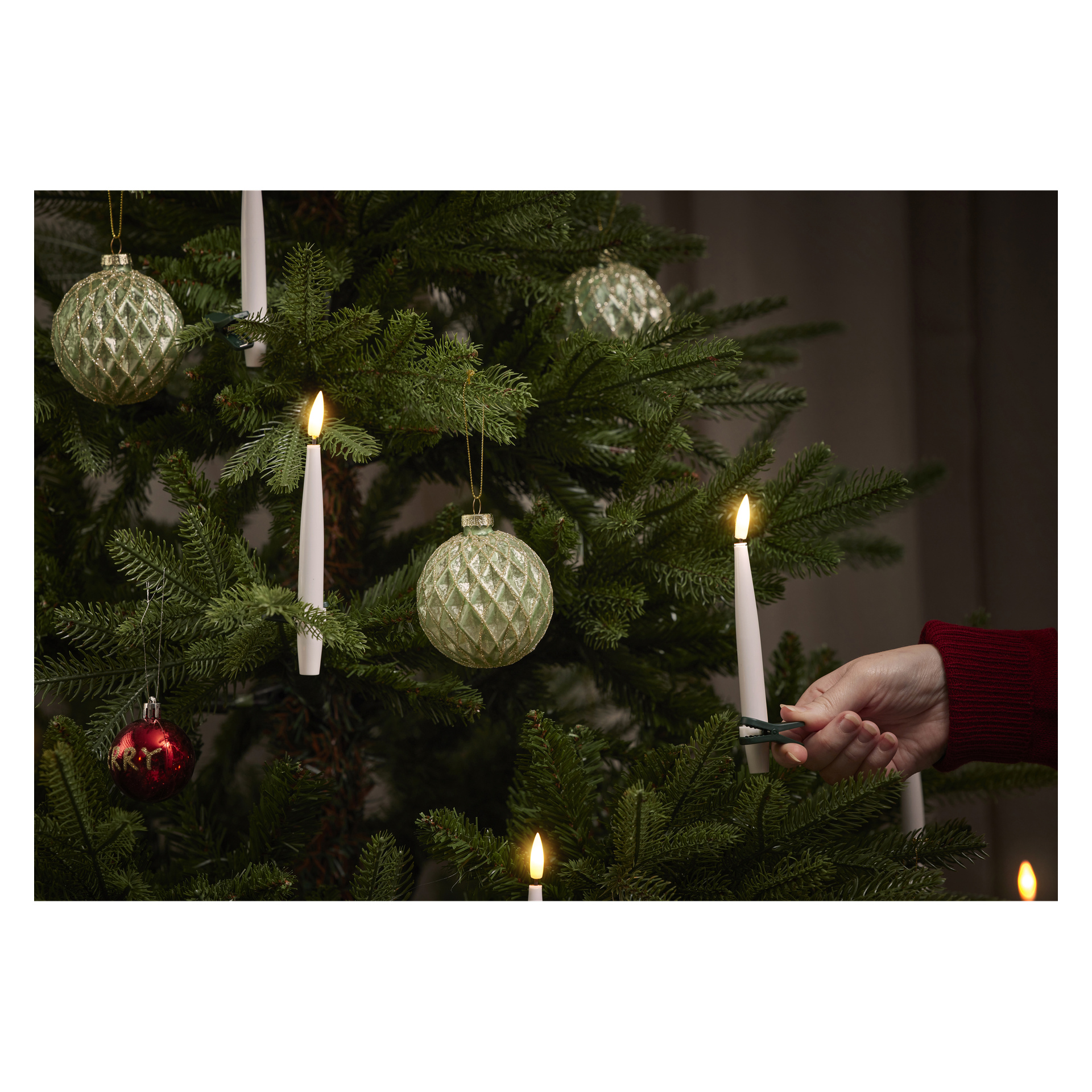 En person som bär röd ärm fäster en EKVIP-batteridriven LED-lampa från en 10-pack till en grön julgran, som pryds med gröna prydnader och en röd prydnad.