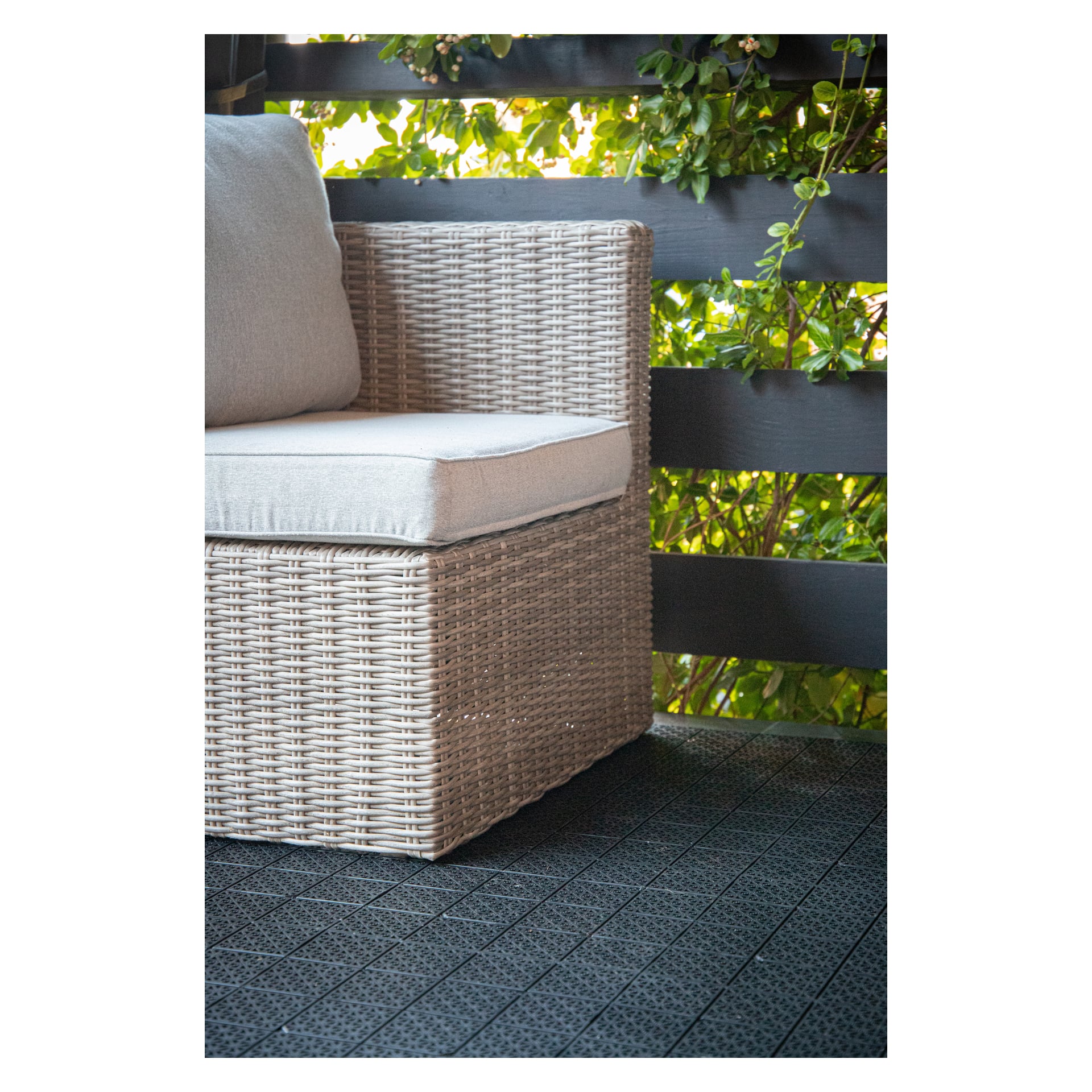 En rottingstol med ljusa kuddar står på ett mörkt kaklat golv gjort med Hard Heads Floor tiles 11-pack. Bakgrunden har ett trästaket och frodiga gröna växter, alla badade i naturligt solljus.