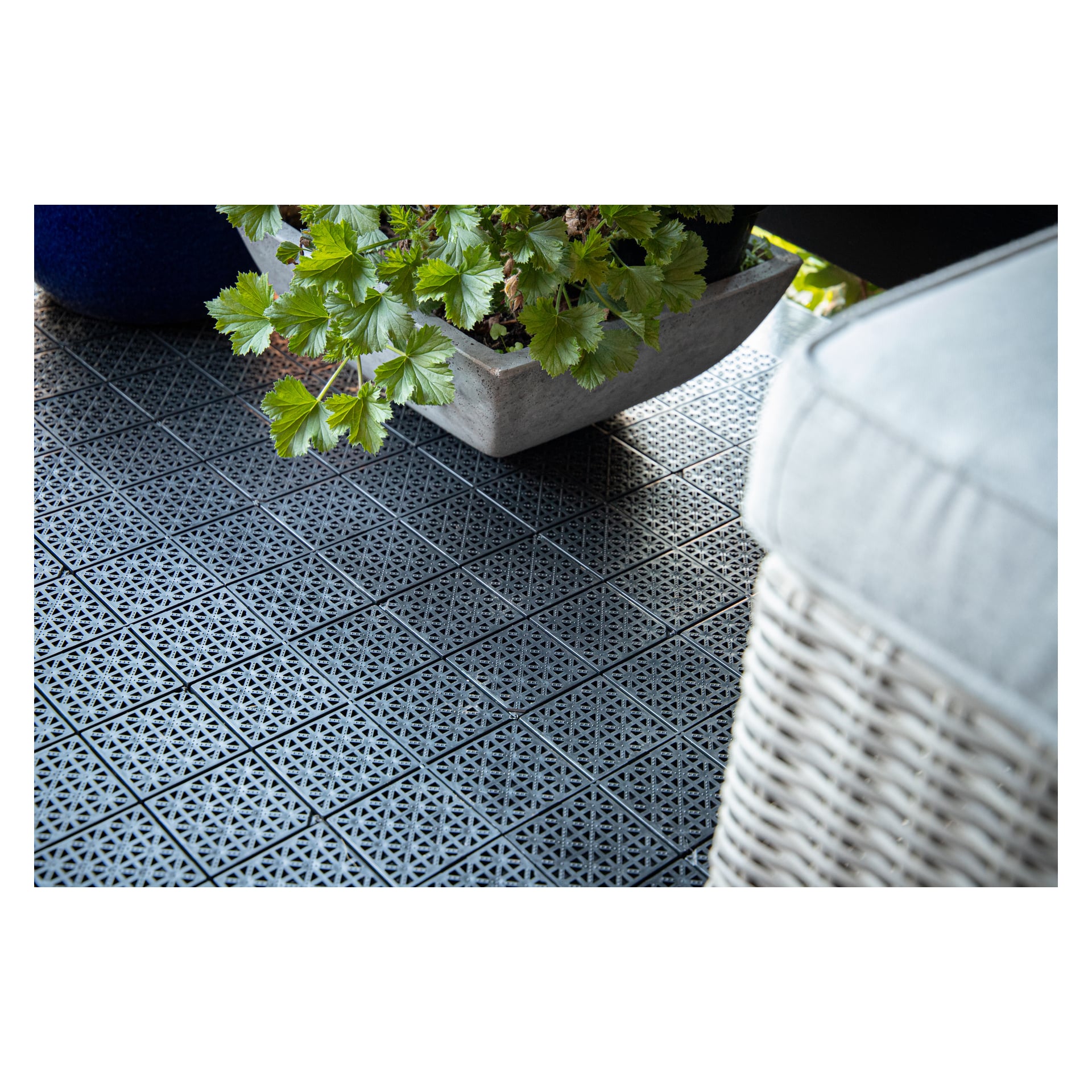 En detaljerad vy av en utomhusyta med Hard Head Floor-plattor i geometrisk design, bredvid en betongkruka fylld med frodigt grönt växtlighet och hörnet av en rottingstol med en blek kudde.