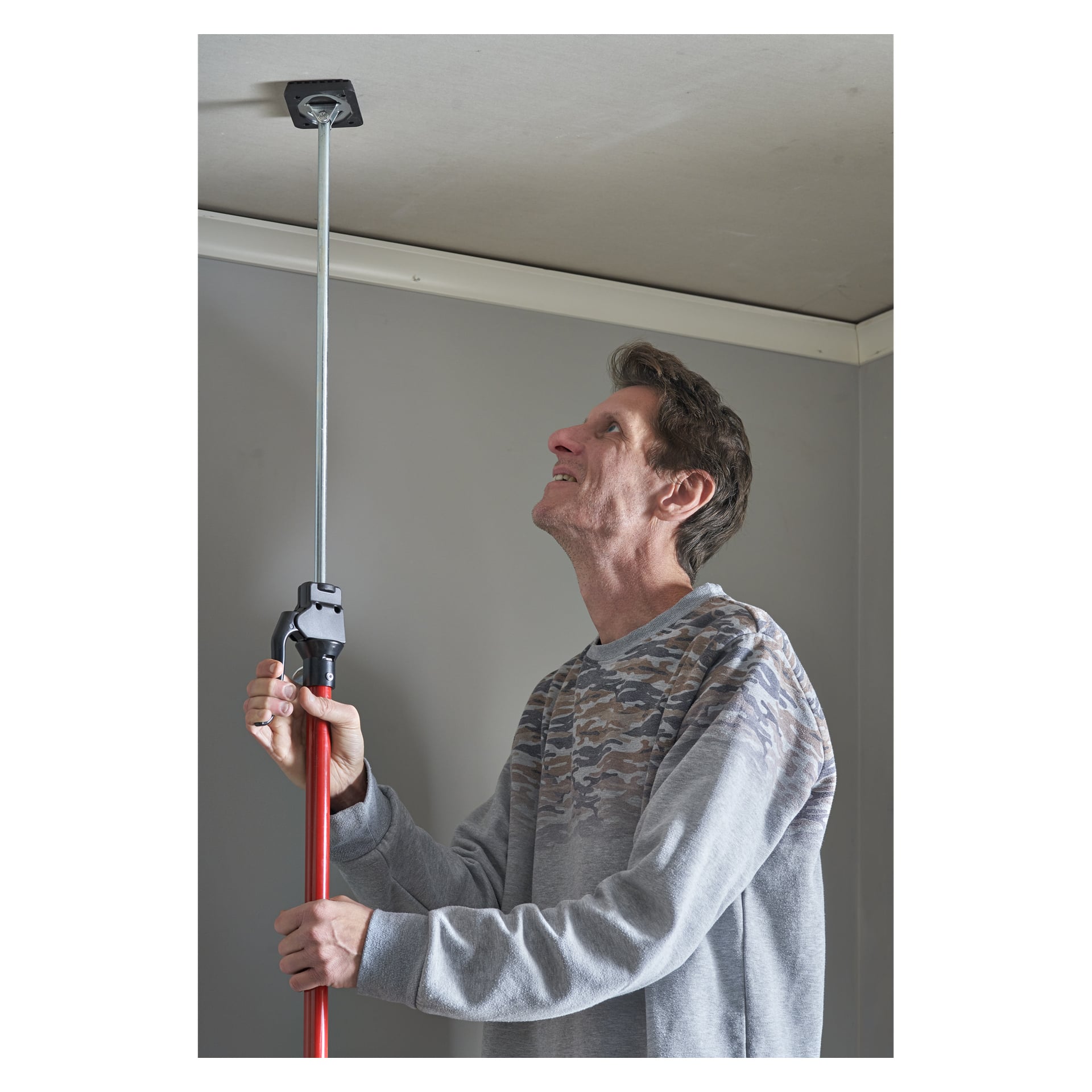 En man i en grå kamouflage-hoodie använder Meec Tools teleskopstöd och trycker upp en fyrkantig platta mot taket i ett rum med grå väggar.