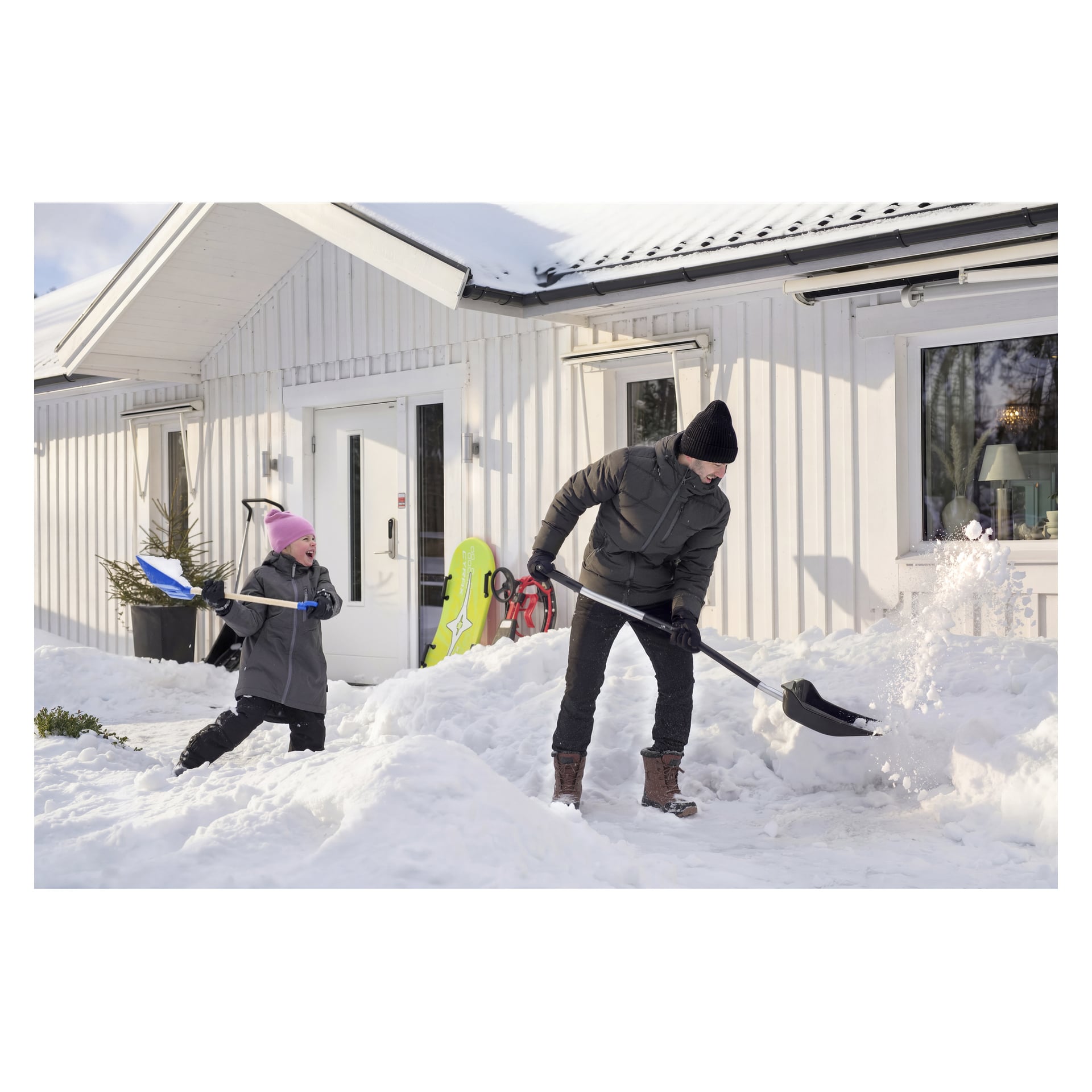 En vuxen och ett barn klar snö med hårda spadar – blå för barnet, svart för den vuxna; Slädarna lutar mot huset.