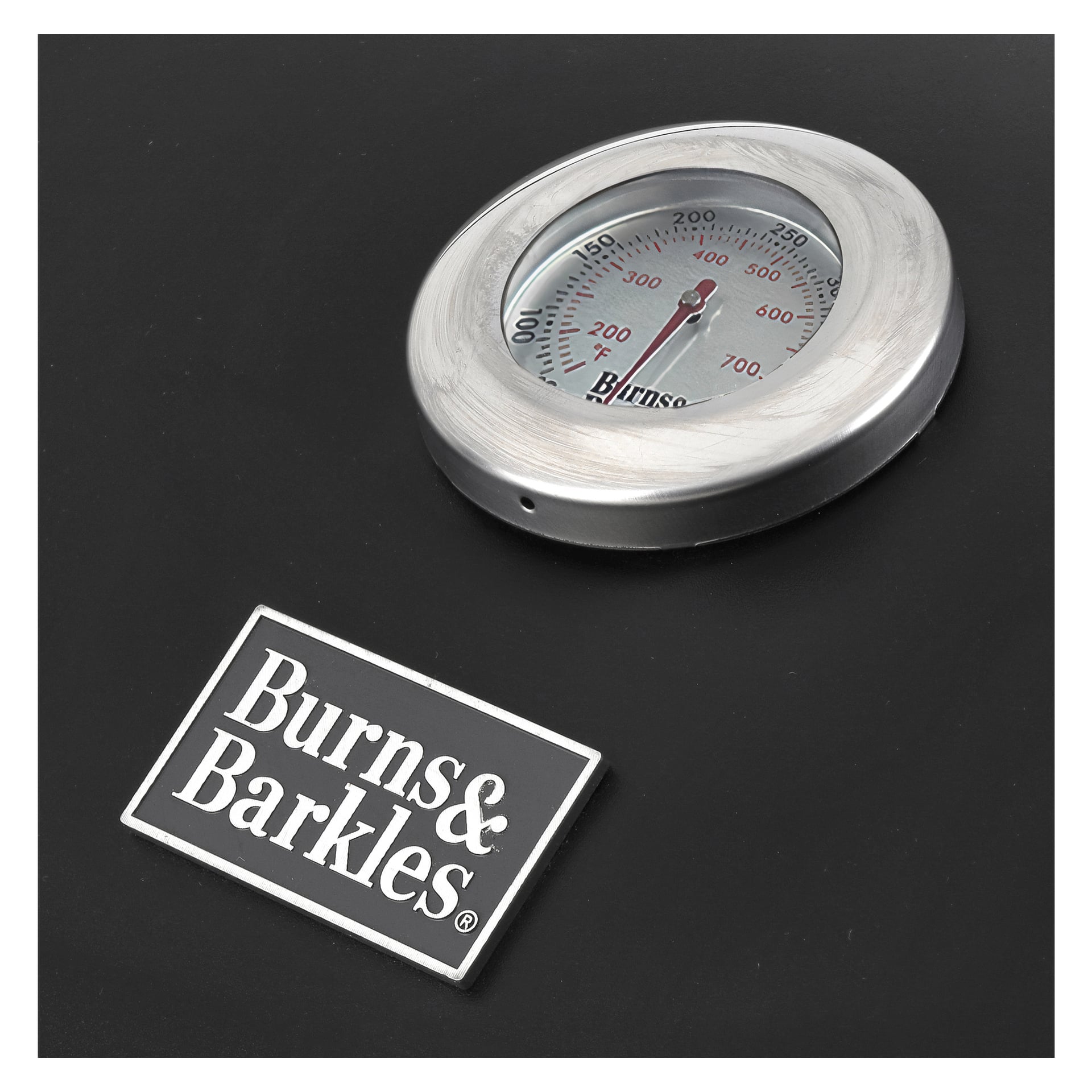 En cirkulär metalltermometer med en röd pekare ligger på en mörk yta bredvid en rektangulär Burns & Barkles-plakett, vilket indikerar dess koppling till den dubbla kolgrillen Coal King 8242. Termometern visar temperaturvärden i Celsius och Fahrenheit.