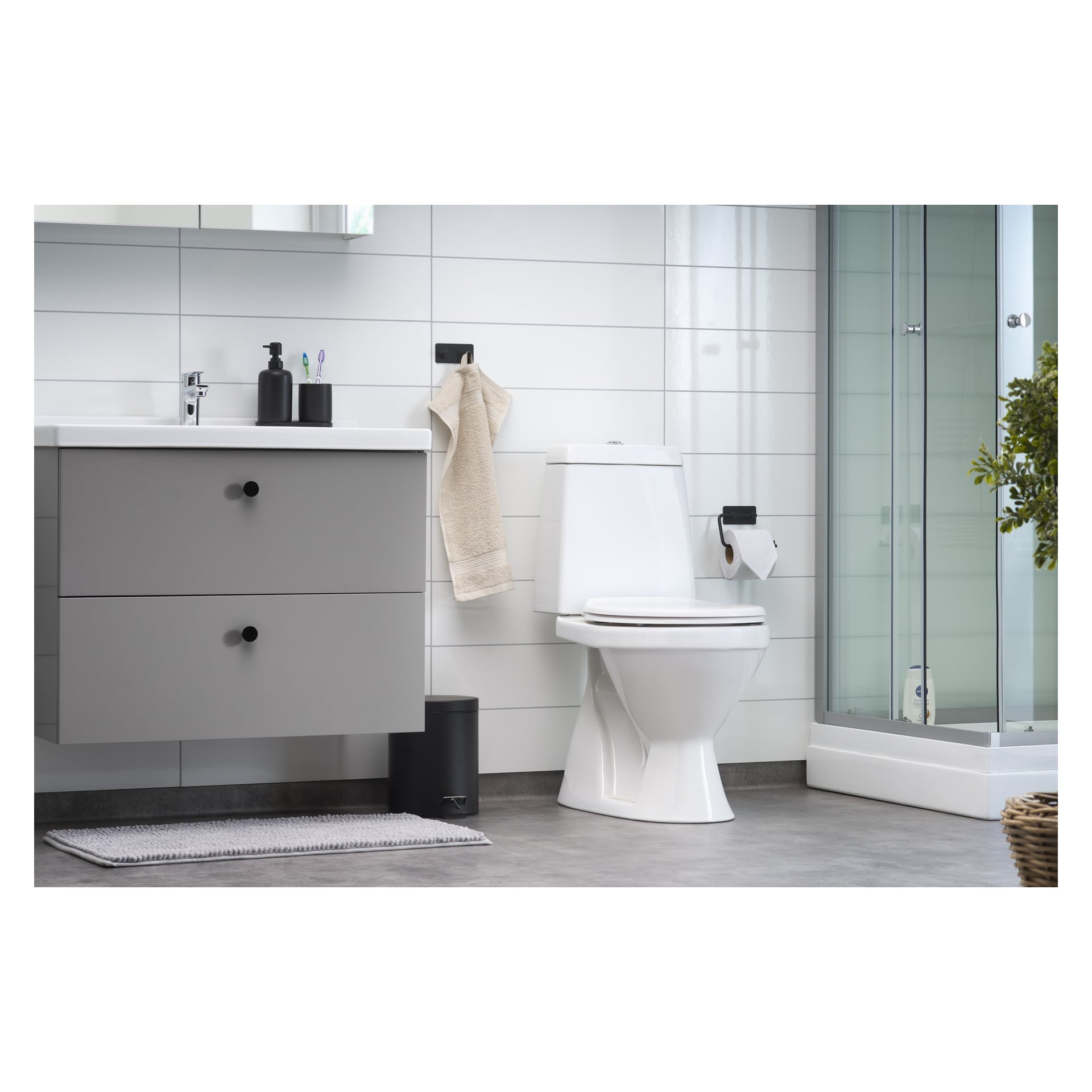 Modern badrum med EKVAP-toaletten, en grå handfatsdisk och en glasdusch. En beige handduk hänger på väggen; Det finns svarta tillbehör, inklusive en tvålbehållare, tandborsthållare och soptunna. En växt är placerad i hörnet.