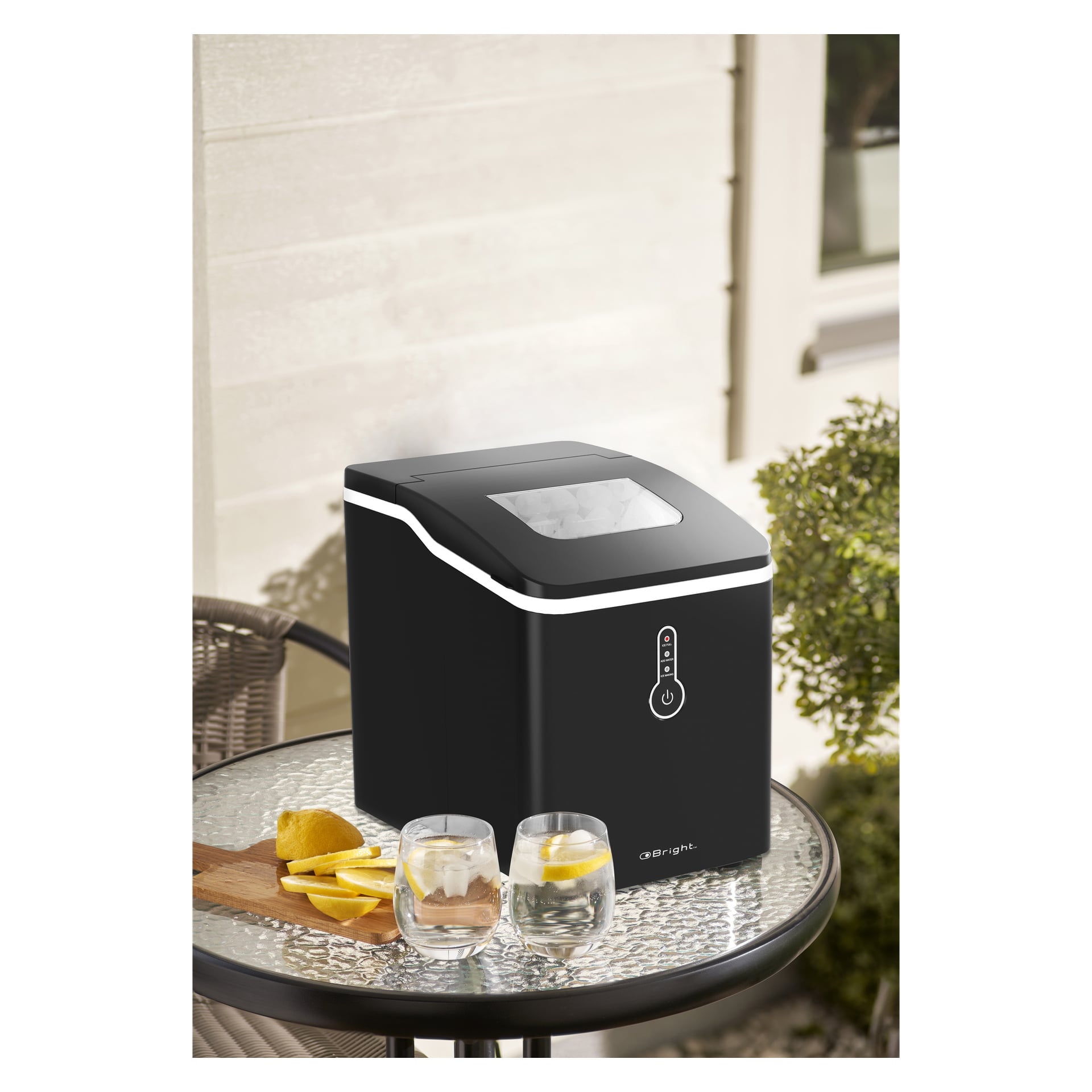 En elegant svart Bright Ice Maker står på ett runt glasbord utomhus, bredvid två glas vatten, citronklyftor och en liten skärbräda i trä. Framsidan har en digital kontrollpanel.