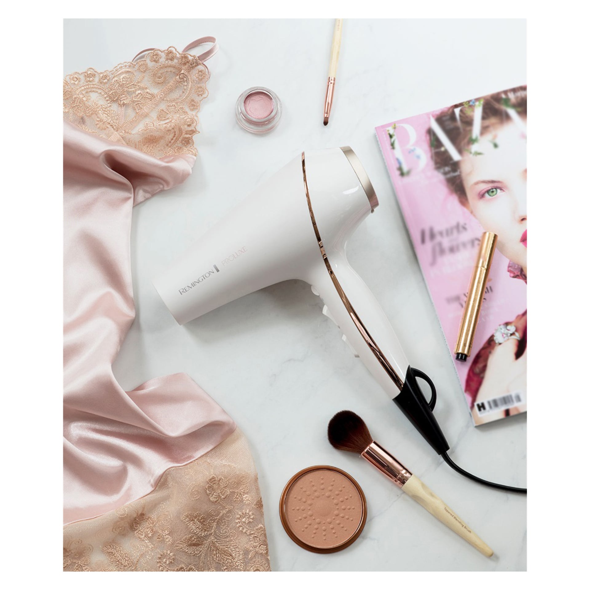 En vit Remington PROLUXE AC9140 hårtork, ett rosa spetsplagg, en sminkborste, en rouge-kompakt, en läppstiftstub, en liten burk smink och ett nummer av Harper's Bazaar är prydligt arrangerade tillsammans.