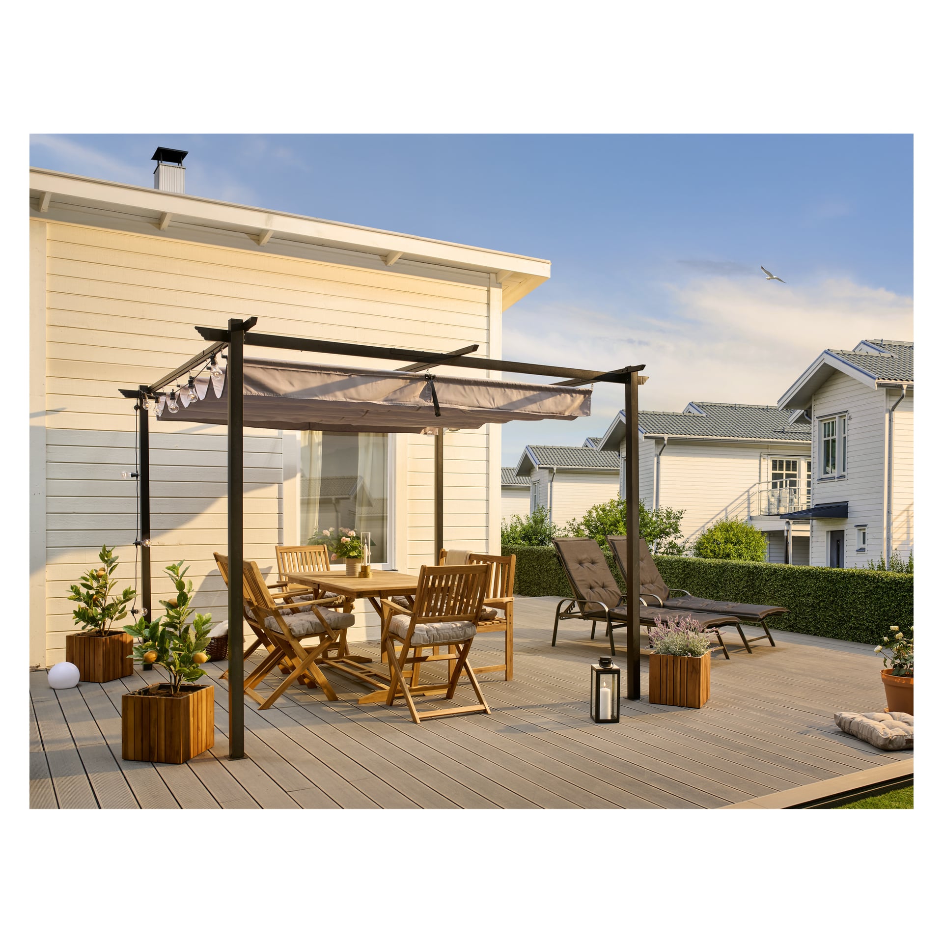En EKVIP Pergola står på en trädäck, med ett matbord och fyra stolar, två solstolar, flera krukväxter och en lykta placerad under den. Däcket gränsar till ett ljust hus, med andra hus i bakgrunden.