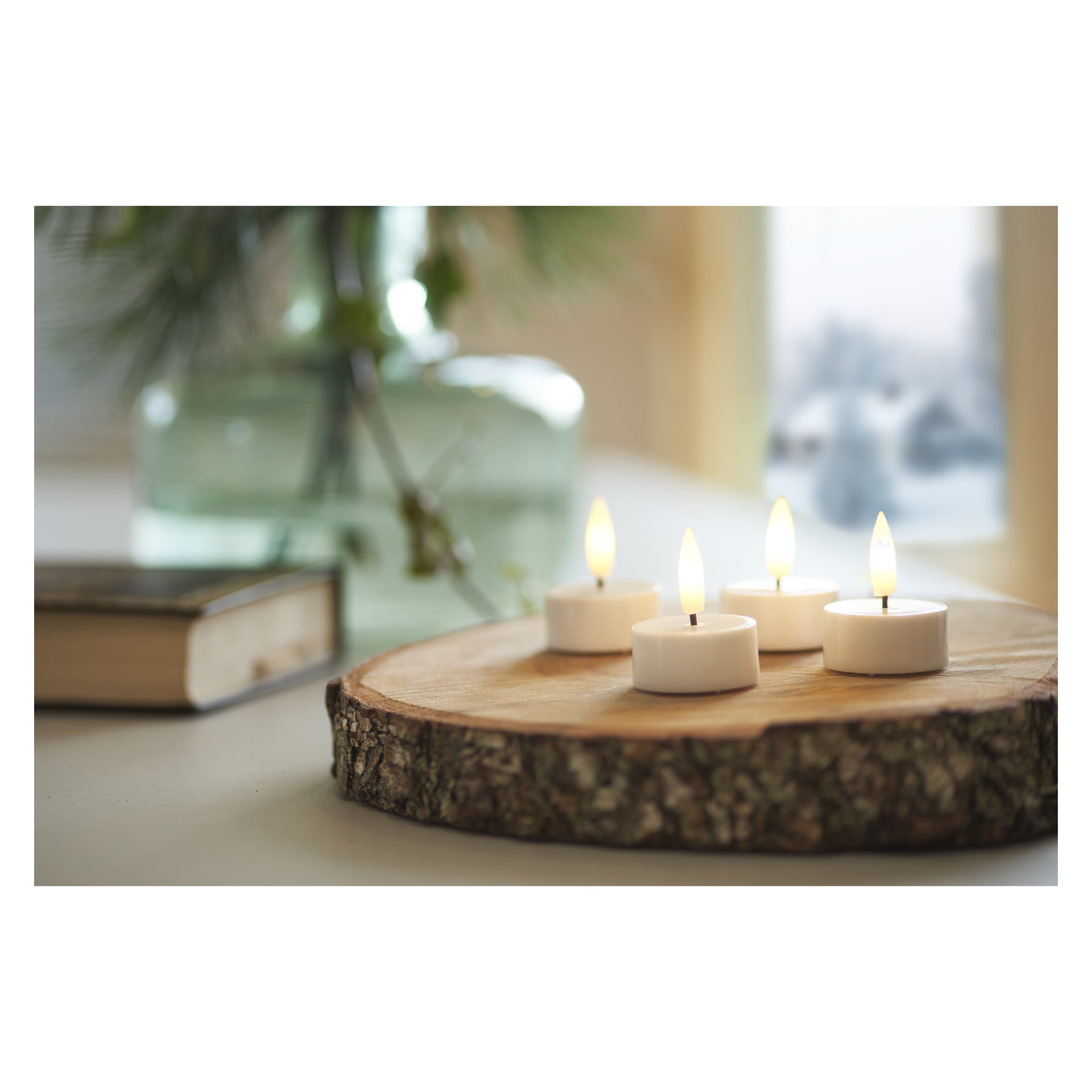 Ett set med fyra upplysta Menuett LED-teljus från ett paket om fyra står på en cirkulär träplatta. Bakom dem finns en stängd bok och en glasvas med grön växtlighet, allt mjukt upplyst av dagsljus som sipprar in genom ett fönster.