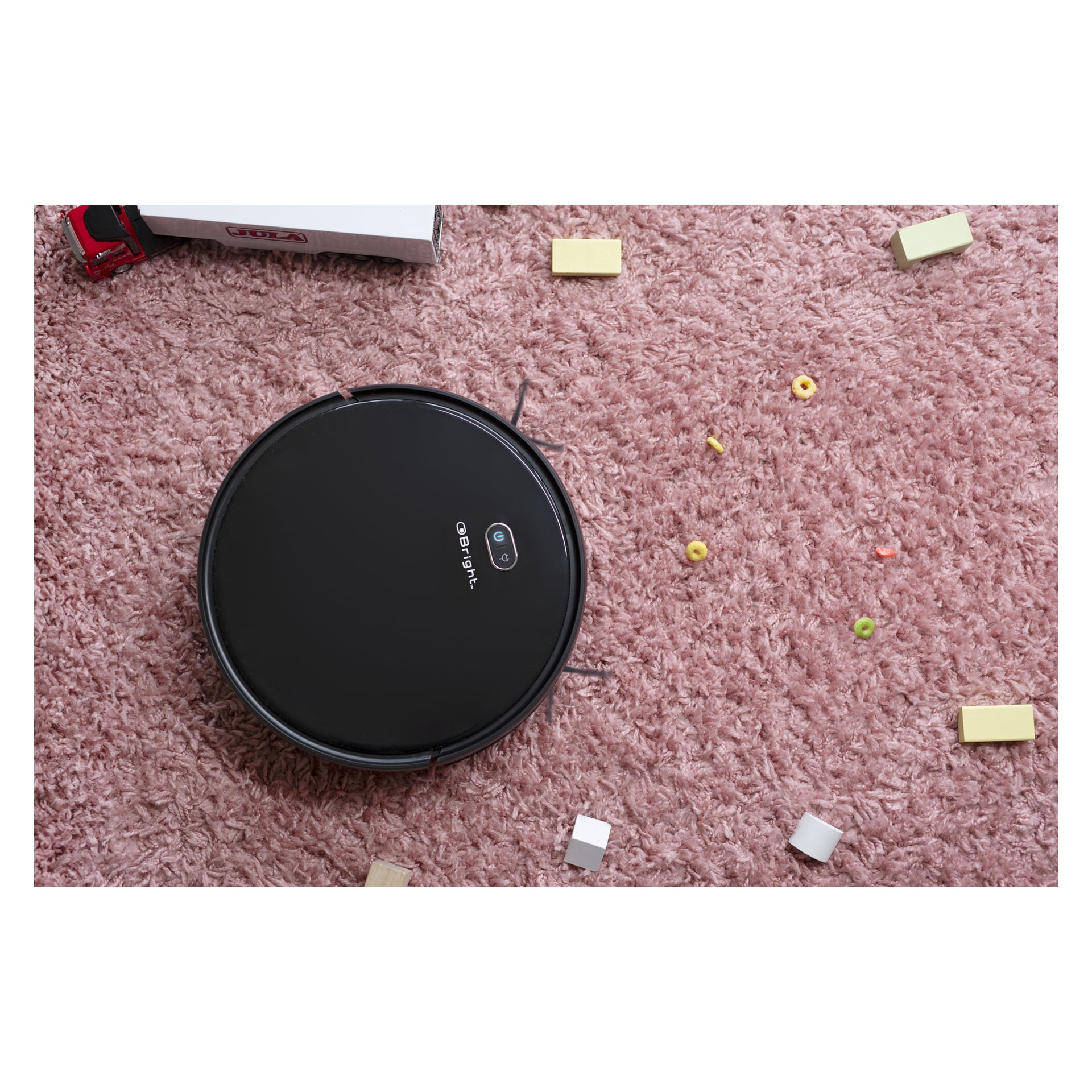 En elegant svart Bright Smart-robotdammsugare rör sig över en rosa matta täckt av leksaker, smulor och träklossar. En leksaksbil syns delvis i det övre vänstra hörnet.