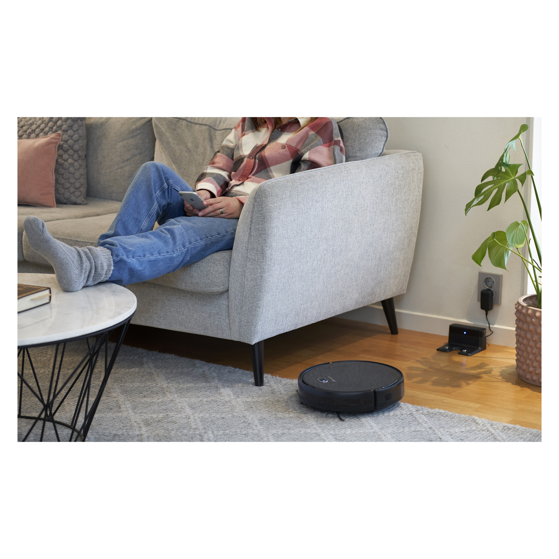 En person slappnar av i en grå soffa inomhus och använder en surfplatta eller telefon medan den svarta Bright Robot-dammsugaren Smart rör sig över det välupplysta trägolvet. I närheten kan man se en laddningsbrygga och en bladväxt.