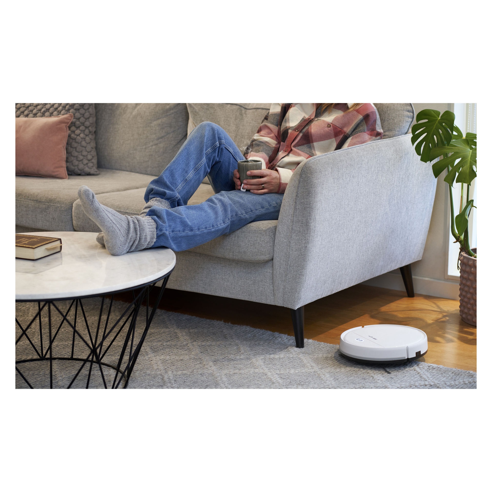 En person slappnar av i en grå soffa med fötterna upp, har strumpor på sig och använder en telefon. I närheten rör sig Bright Robotic-dammsugaren över trägolvet bredvid en matta och ett soffbord fyllt med böcker.