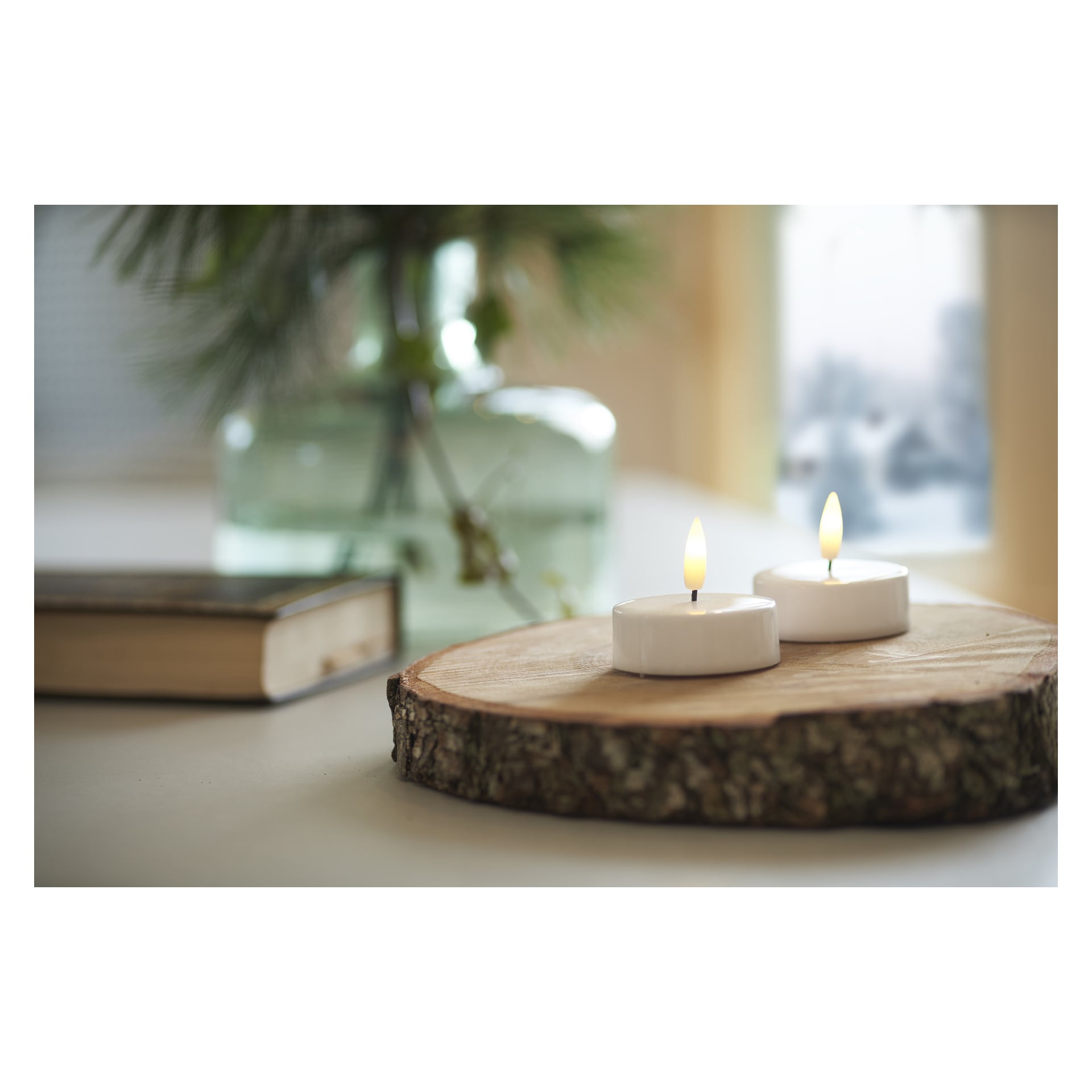 Ett par Menuett Large Tea Lights med fladdrande LED-lågor vilar på en cirkulär träplatta. Bakom dem dyker en stängd bok och en glasvas fylld med gröna blad mjukt ur fokus, badande i milt dagsljus som strömmar från ett närliggande fönster.