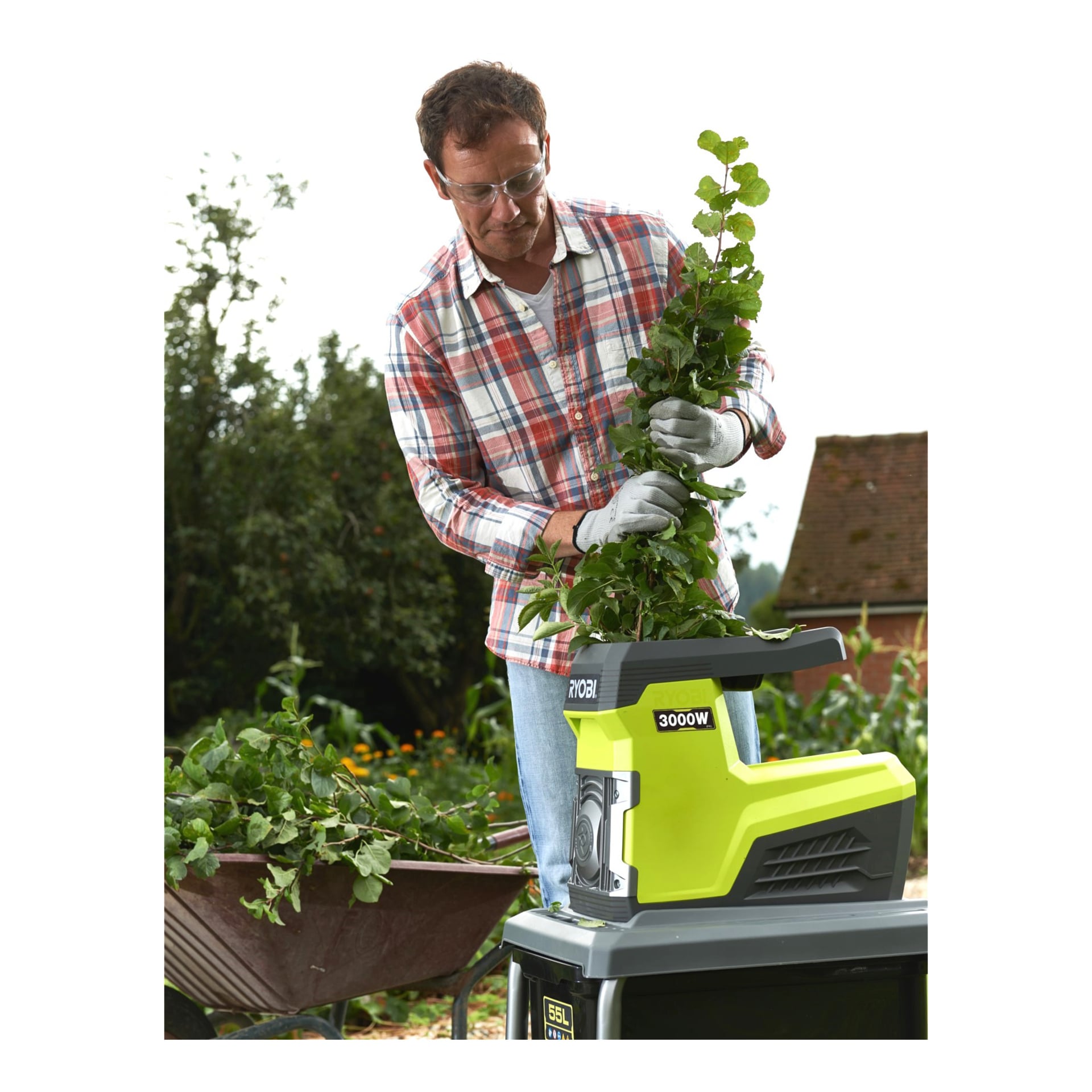 En man med glasögon och handskar är utomhus och matar grenar i en Ryobi Compost-strimlare RSH3045U. En skottkärra fylld med avskurna grenar står i närheten, och träd syns bakom honom.