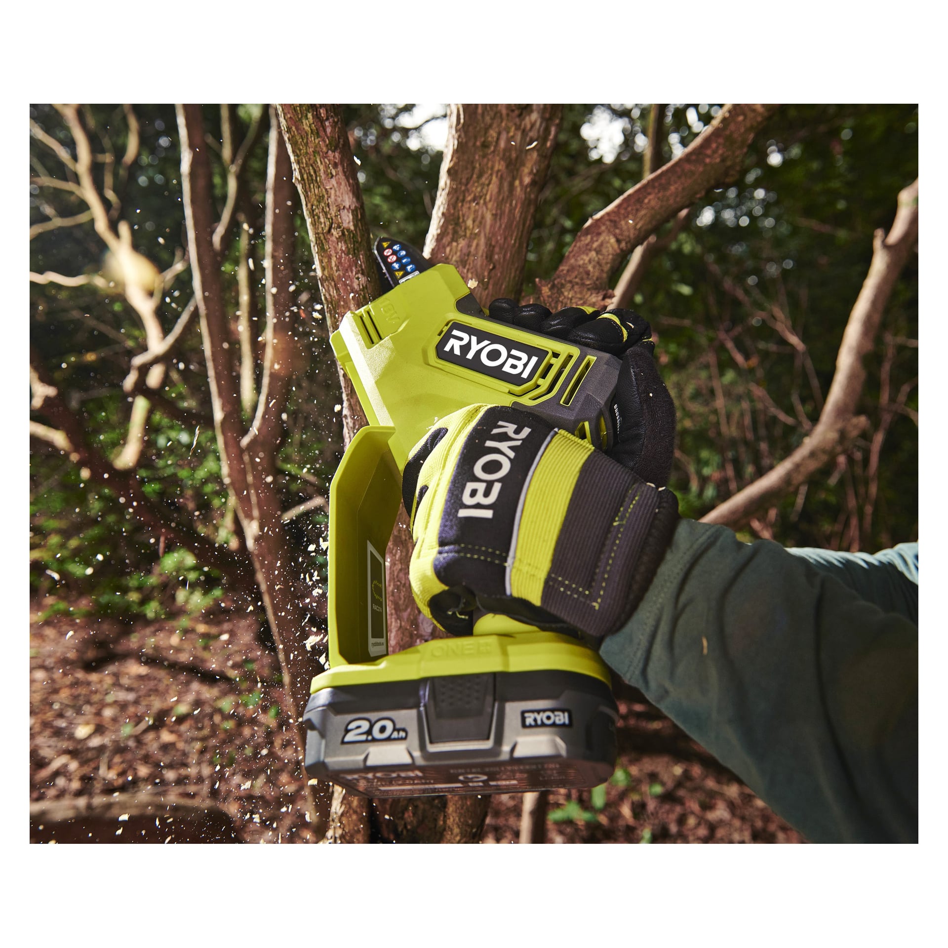 Någon i svarta och gröna handskar och en mörk jacka använder en Ryobi One+ RY18PSX10A-120 batteridriven beskärningssåg för att klippa en trädgren utomhus, medan träflisor sprids i luften.