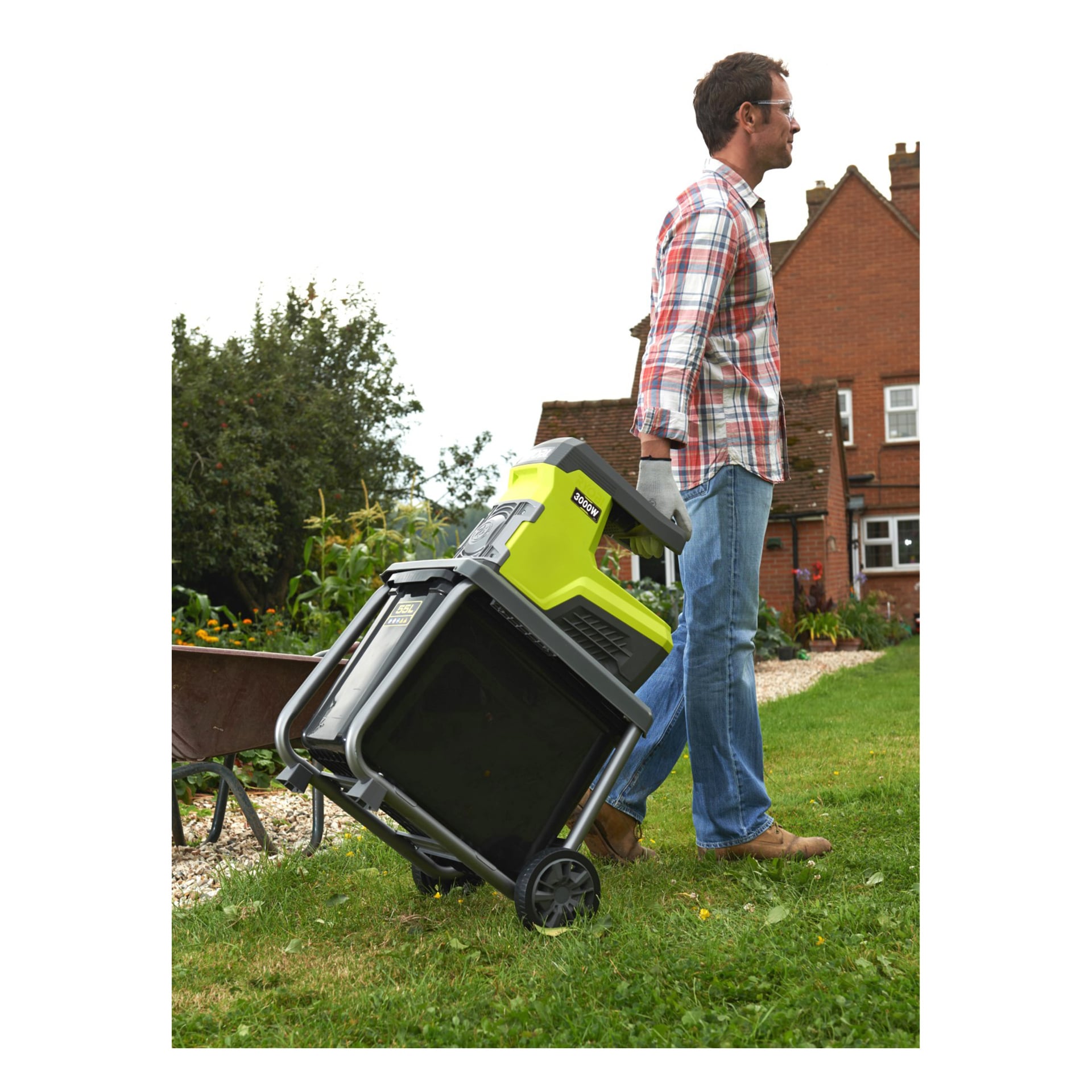 En man i rutig skjorta och jeans drar en grön och svart Ryobi Compost Shredder RSH3045U på hjul genom en gräsmatta, med ett tegelhus och en trädgård synliga bakom sig.