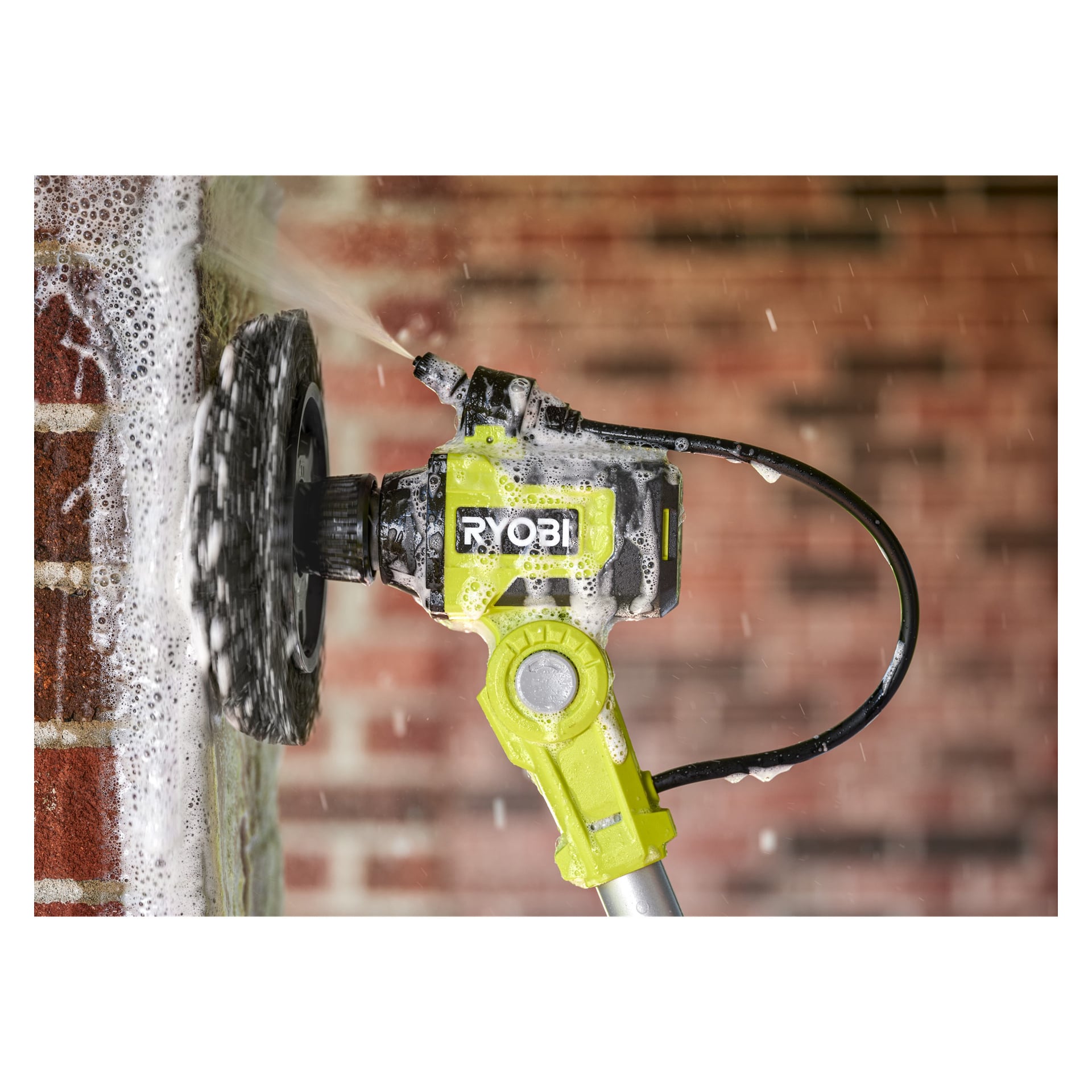 Szczotka Ryobi One+ Ring RACLTH18, nałożona mydłem, szoruje ceglaną ścianę i generuje piankowy spray. Tło widać delikatnie rozmazane czerwone cegły.