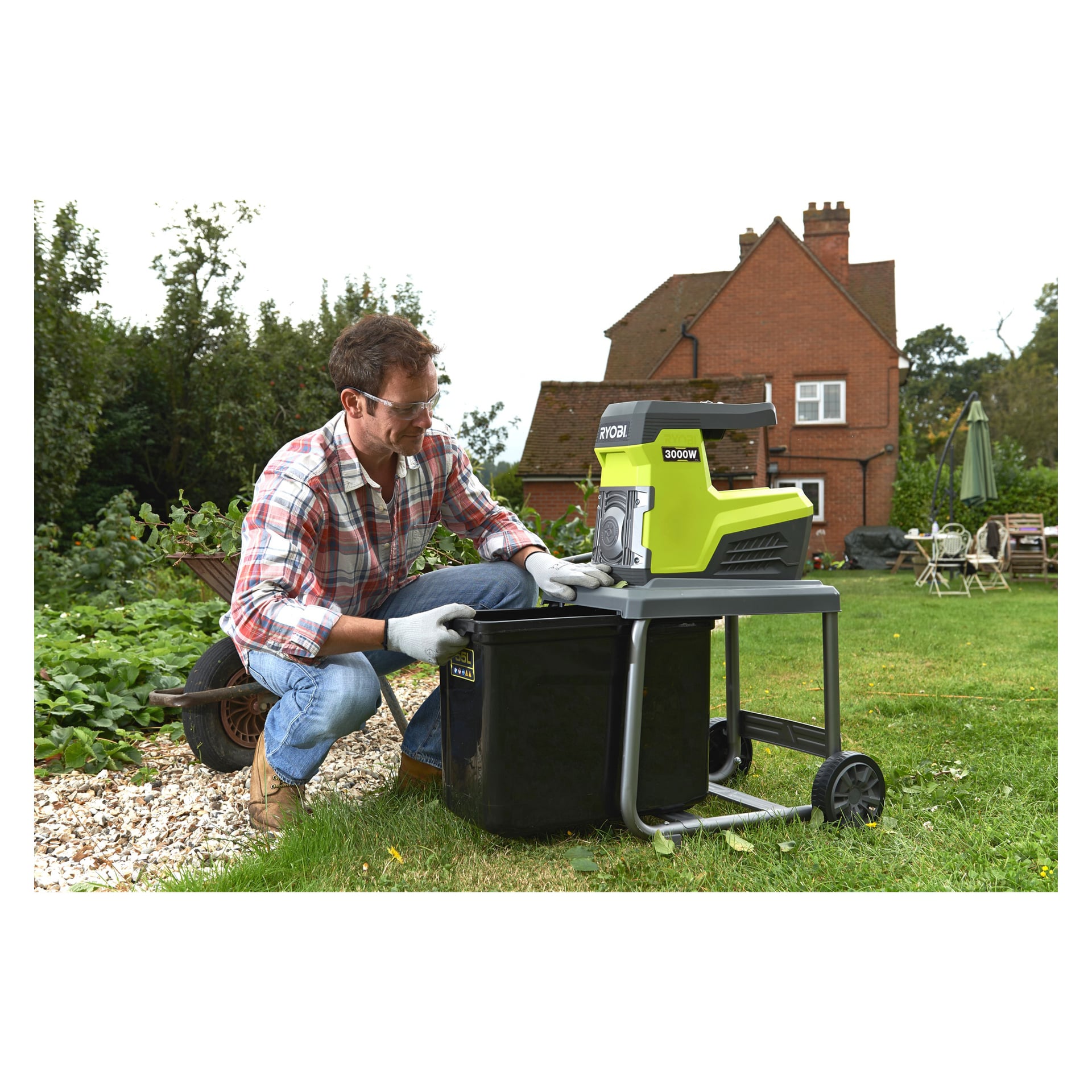 En man med skyddsglasögon och handskar knäböjer på gräset bredvid en Ryobi Compost Shredder RSH3045U och lägger skräp i en svart soptunna. I bakgrunden finns ett hus, utomhussittplatser och frodig grönska.