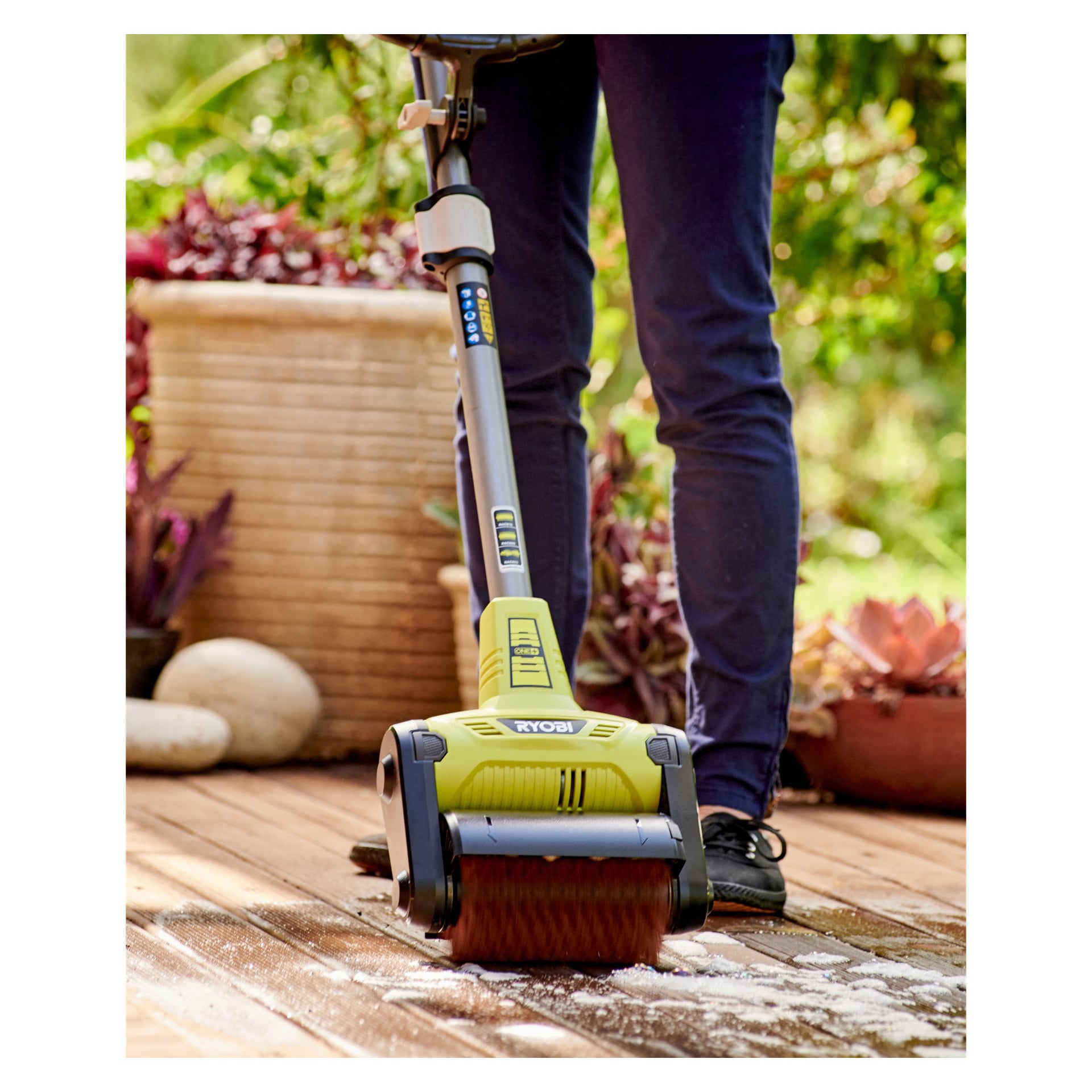 En Ryobi One+ skrubbborste används av någon för att rengöra en trädäck och skjuta bort vitt skräp. Scenen inkluderar krukväxter i bakgrunden, och endast individens ben och fötter syns.