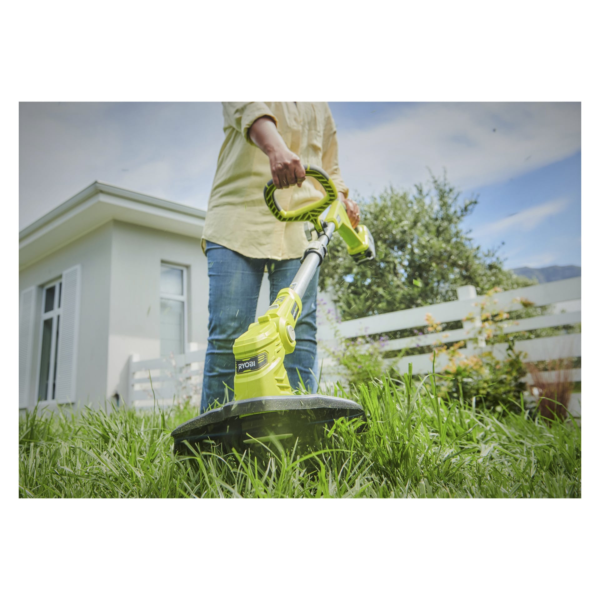 Man klipper gräs med en Ryobi One+ batteridriven trimmer OLT1832A i en solig trädgård, med ett vitt hus, staket och trädgårdsväxter synliga bakom sig.