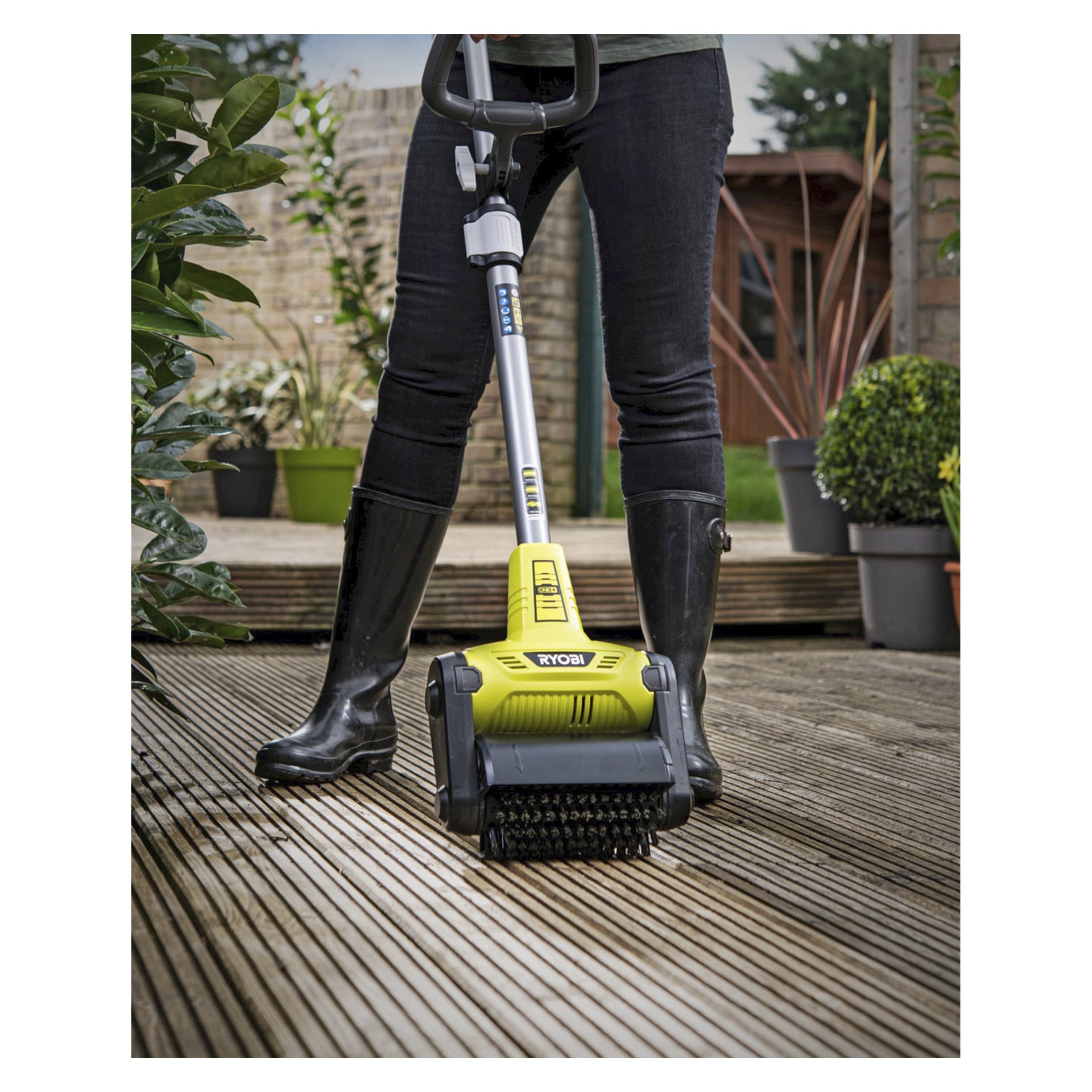 En person i svarta stövlar kör den gula Ryobi One+ Patio cleaner RY18PCB-0 på en utomhusaltan i trä, omgiven av krukväxter och med ett trädgårdsskjul i bakgrunden.