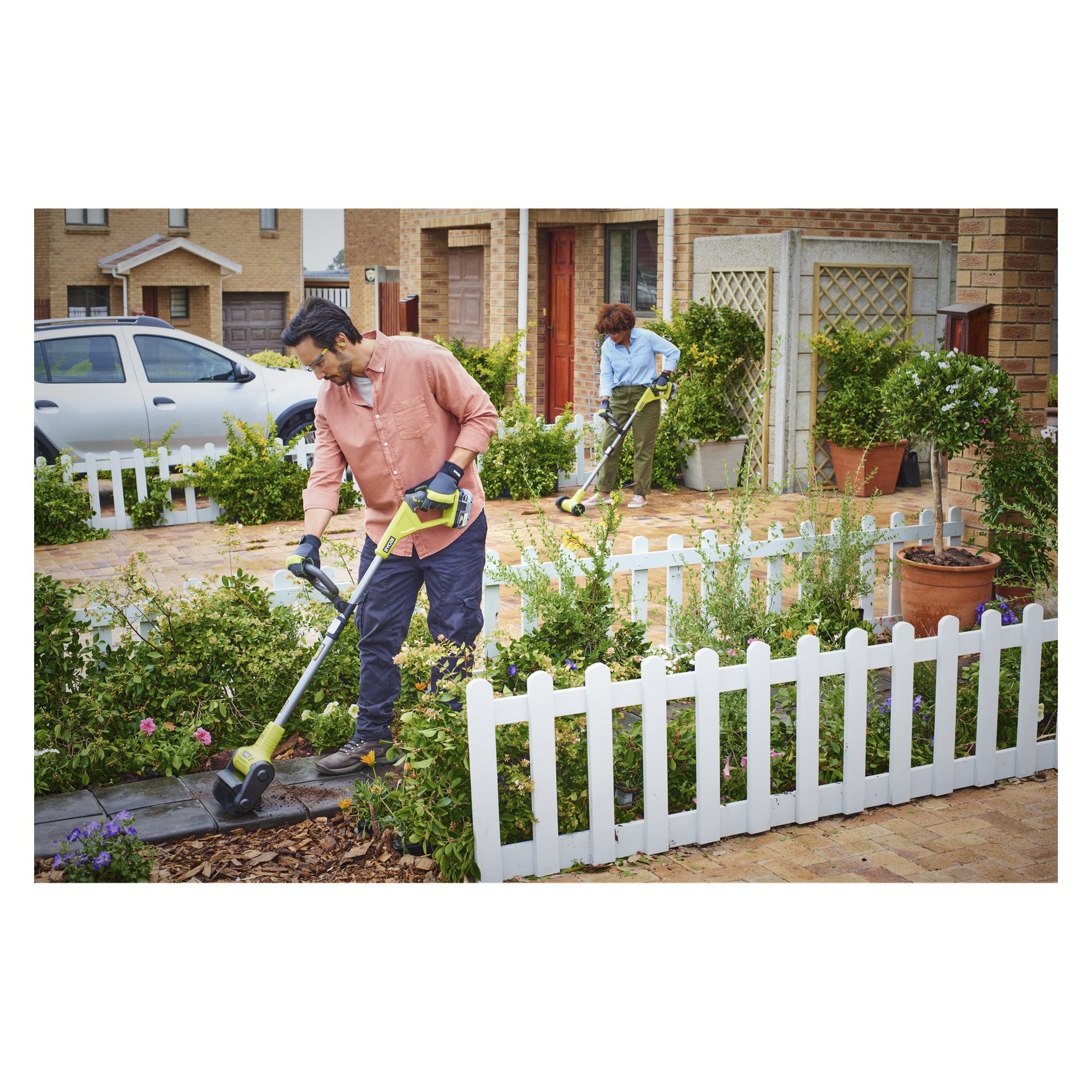 En man driver en Ryobi One+ Patio cleaner RY18PCB-0 på en trädgårdsgång, medan en kvinna i närheten klipper gräs nära ett vitt staket i en förortsträdgård. Det finns olika buskar och blommande växter runtomkring, och en bil står parkerad på uppfarten.