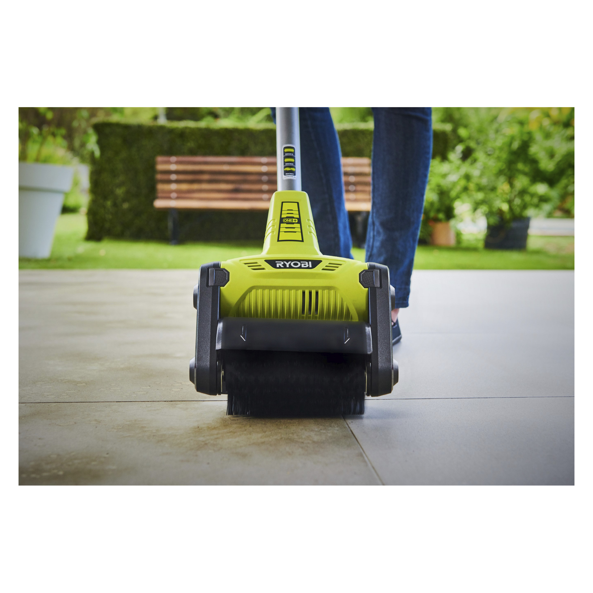 En person använder en grön Ryobi One+ Patio Cleaner RY18PCB-0 för att skrubba en kaklad uteplats, med en träbänk, frodiga växter och grönska som mjukt är suddig i bakgrunden.