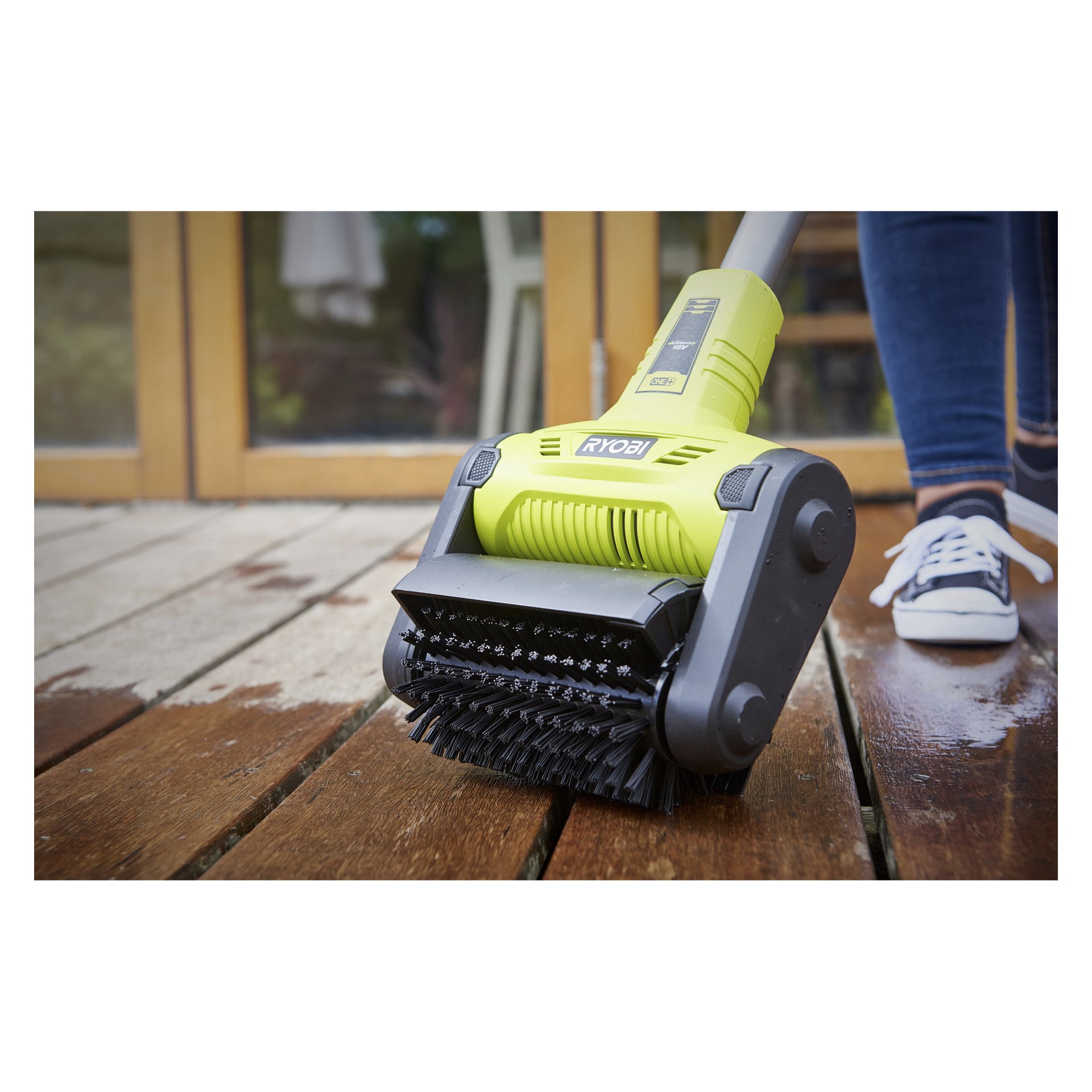 Någon rengör en våt träaltan med en Ryobi One+ Patio cleaner RY18PCB-0, som har roterande borstar. Personen är klädd i jeans och svartvita sneakers.