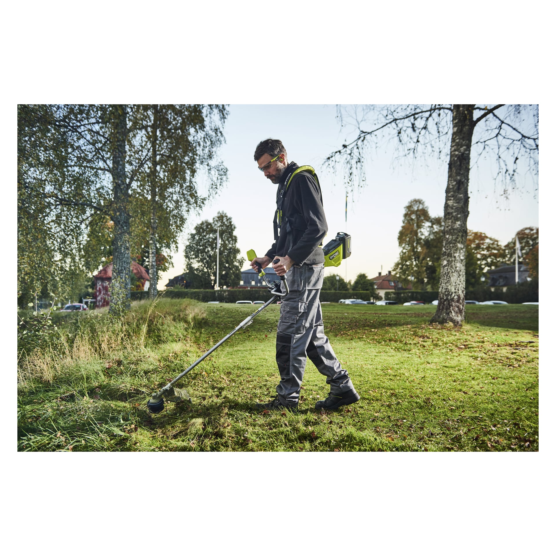 Med skyddsglasögon och arbetskläder klipper man gräs med en sladdlös trimmer i en park med träd och hus i närheten.