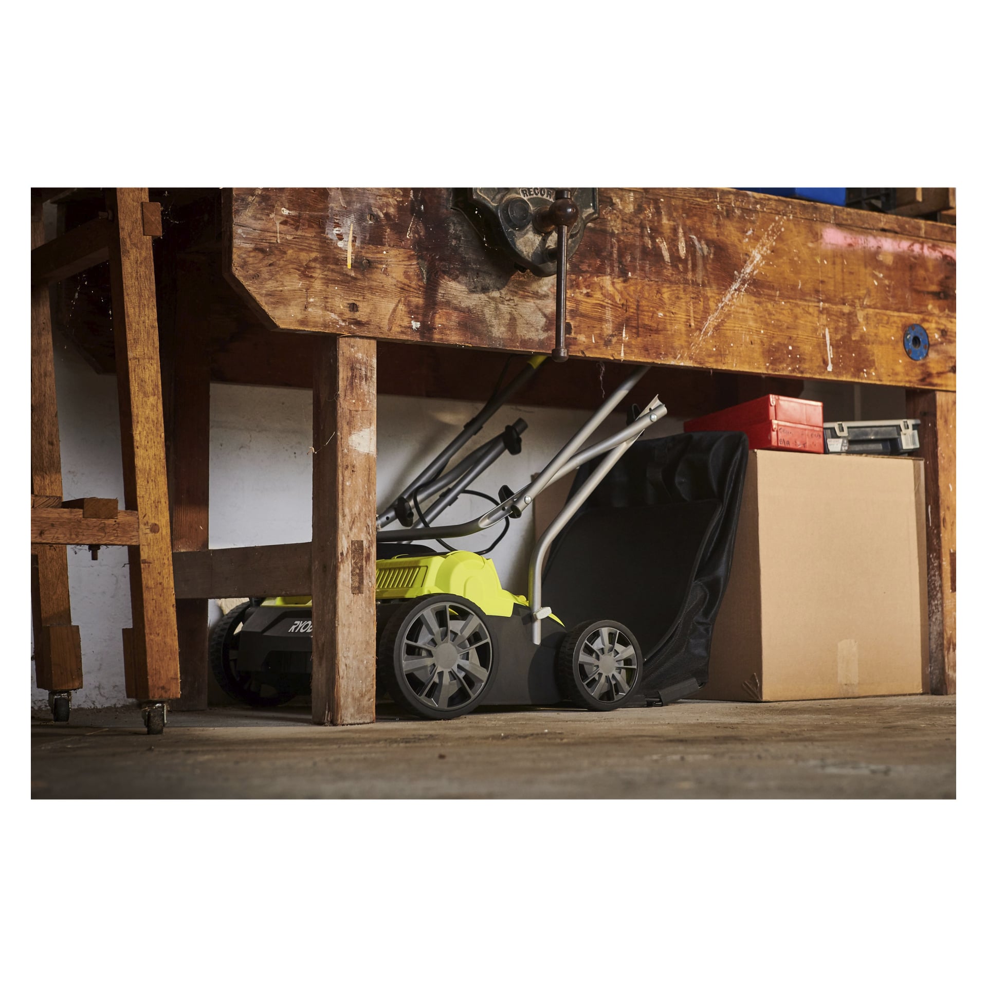 En gul och svart Ryobi One+ batteridriven verticutter RY18SFX35A-0 är placerad under en träarbetsbänk. Nära står en kartong, en röd verktygslåda och ett svart plastfodral, vilket indikerar att området troligen används som garage eller verkstad.