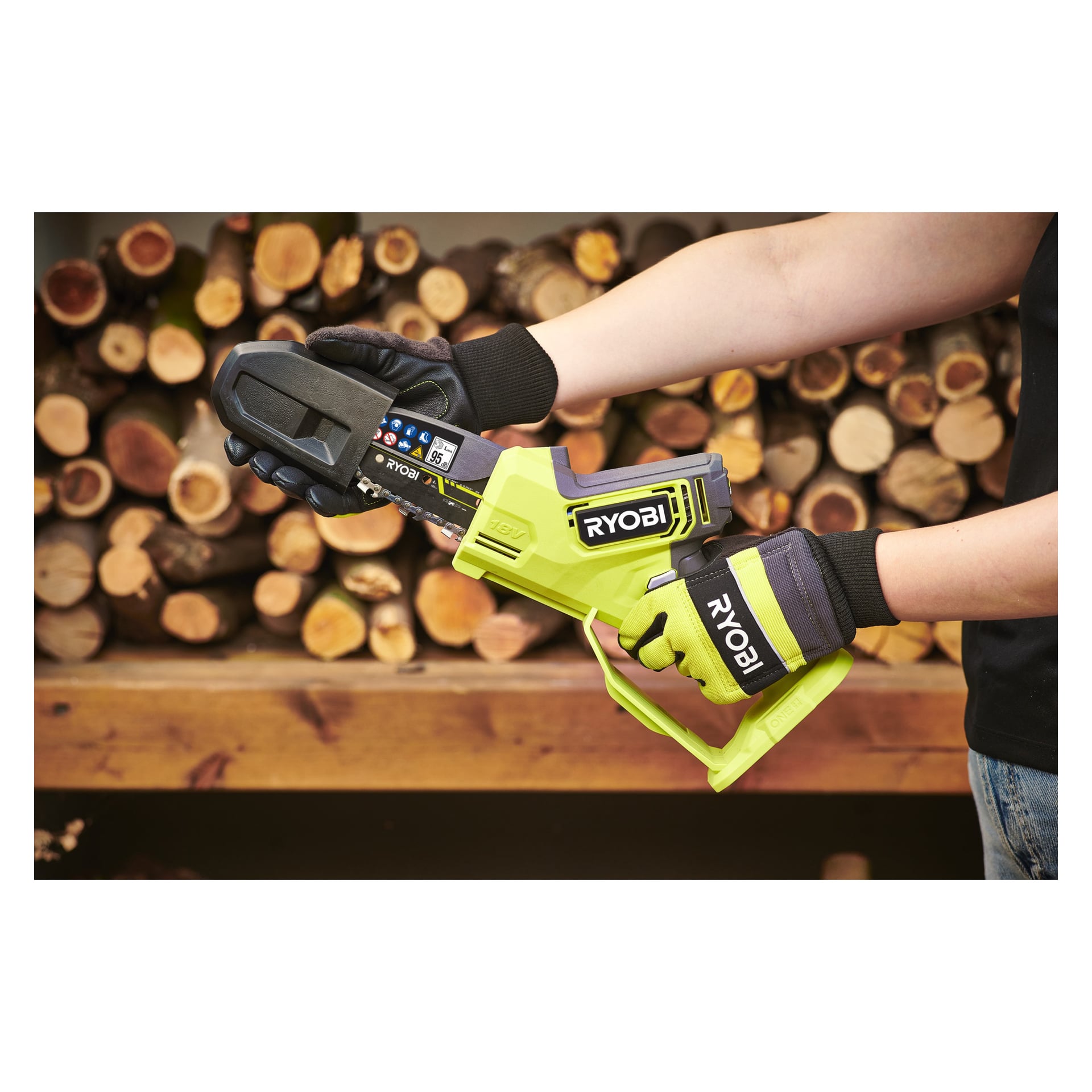 En person med svarta handskar håller i en grön Ryobi One+ batteridriven beskärarsåg, modell RY18PSX10A-120, och håller den horisontellt framför en hög med vedstockar. Endast deras handskbeklädda händer och en del av armen visas, redo att hantera verktyget.