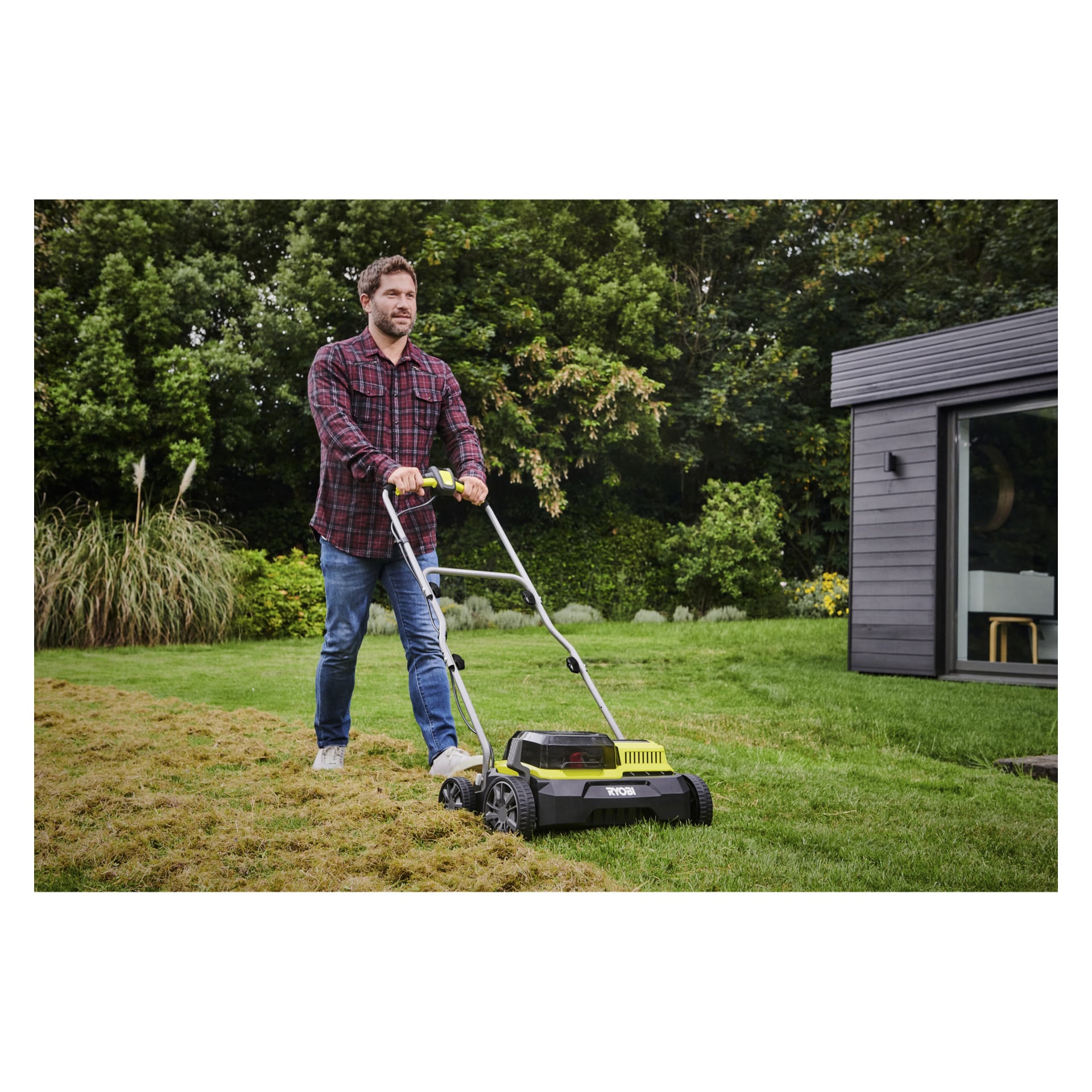 En man klädd i en röd rutig skjorta och jeans kör Ryobi One+ Battery-drivna verticutter RY18SFX35A-0 på en grön gräsmatta bredvid ett modernt mörkt hus, med frodig växtlighet runtomkring.