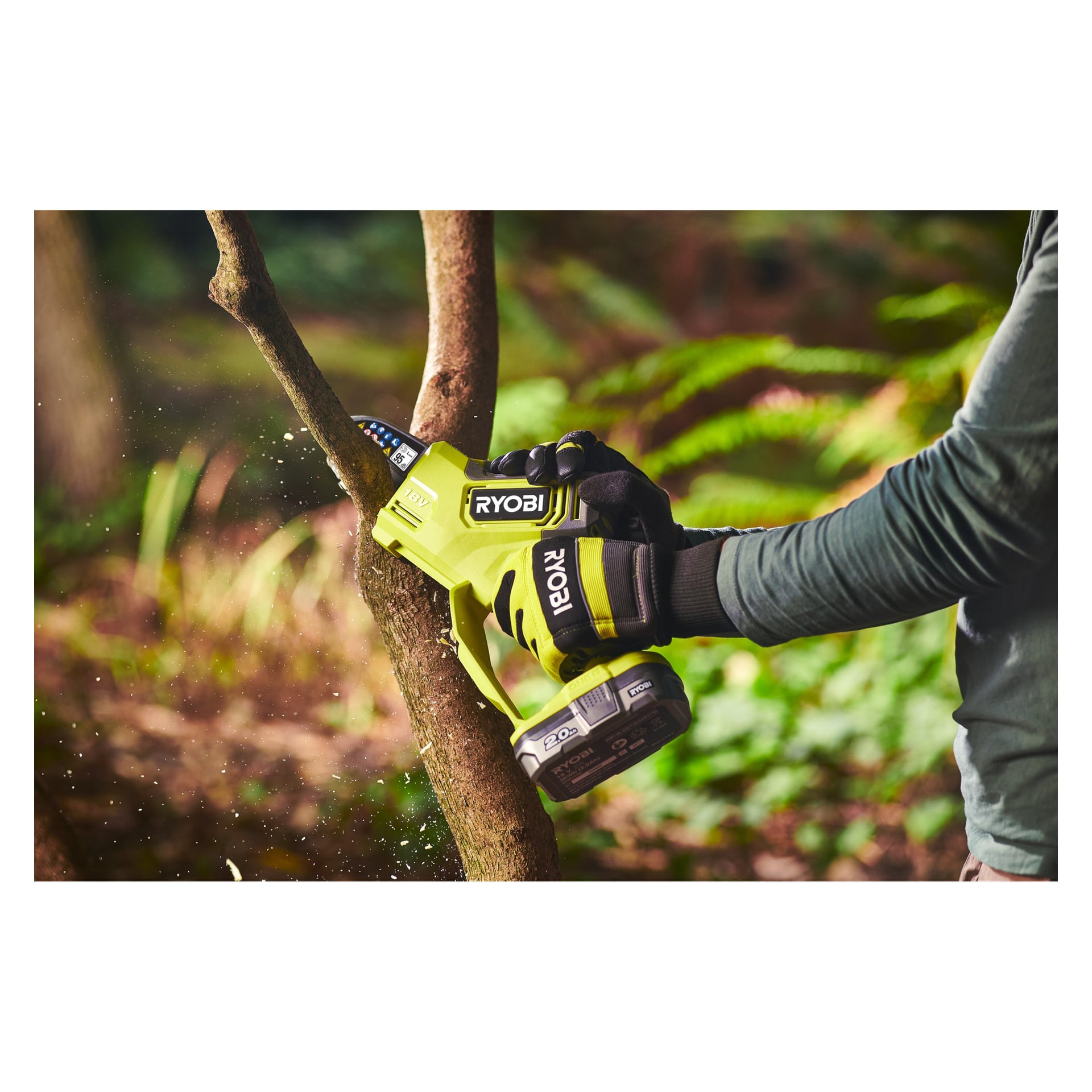 En person i svarta handskar och långärmad skjorta använder en Ryobi One+ batteridriven beskärningssåg RY18PSX10A-120 för att trimma en utomhusgren, vilket skickar träflis upp i luften när bladet skär igenom.
