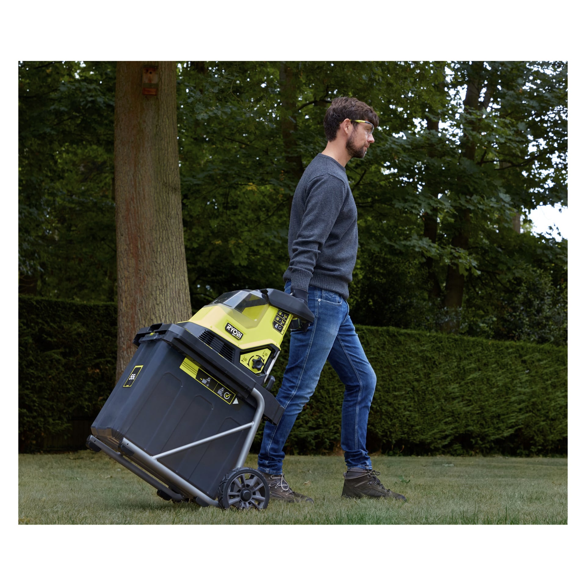 En man i grå tröja och jeans drar en gul och svart Ryobi Max Power batteridriven kompoststrimler RY36SHX40-0 med hjul över en gräsmatta, med träd och en häck synliga bakom sig.