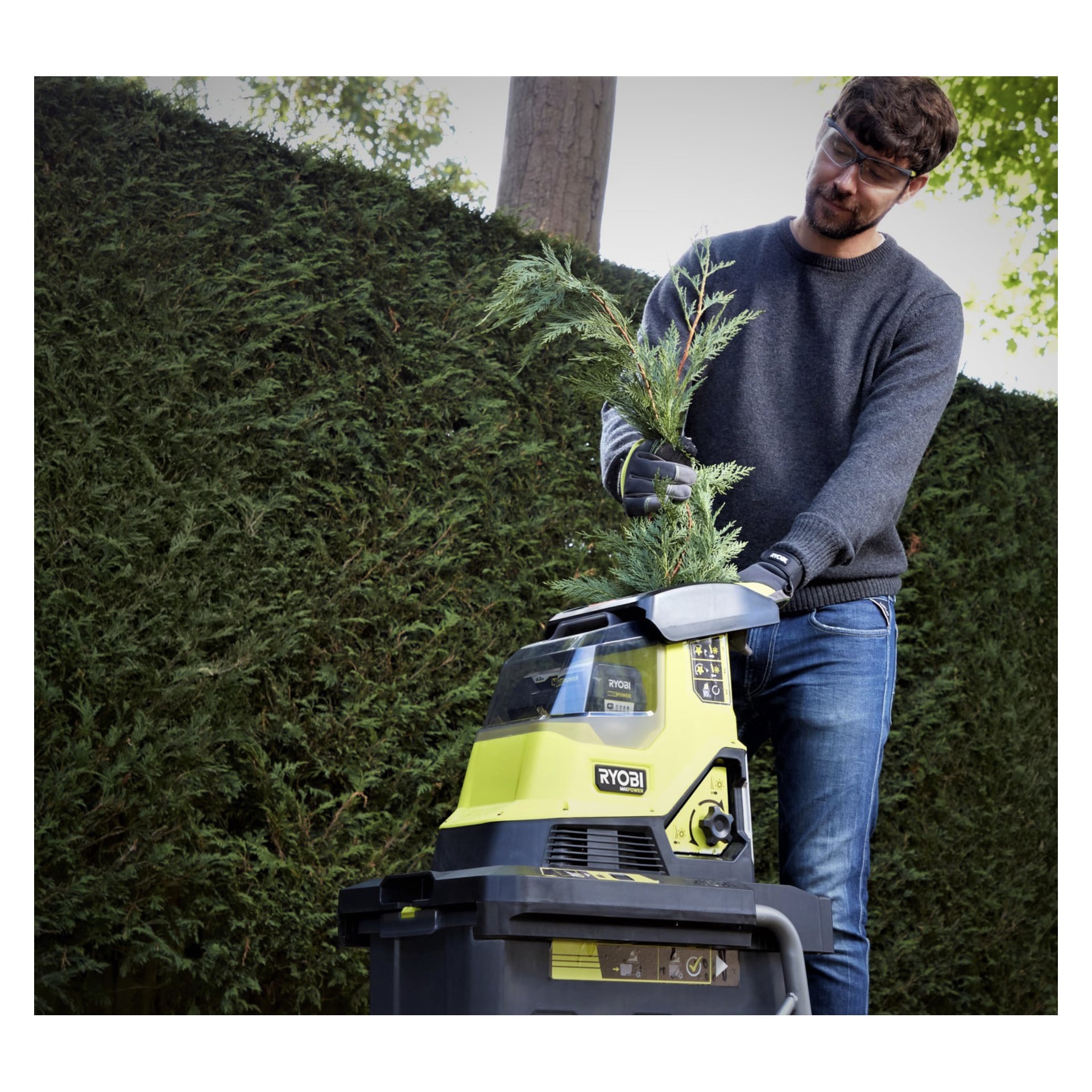 En person med skyddsglasögon och handskar lägger en gren i en Ryobi Max Power batteridriven kompoststrimlar (modell RY36SHX40-0) utomhus, med en hög häck synlig bakom dem.