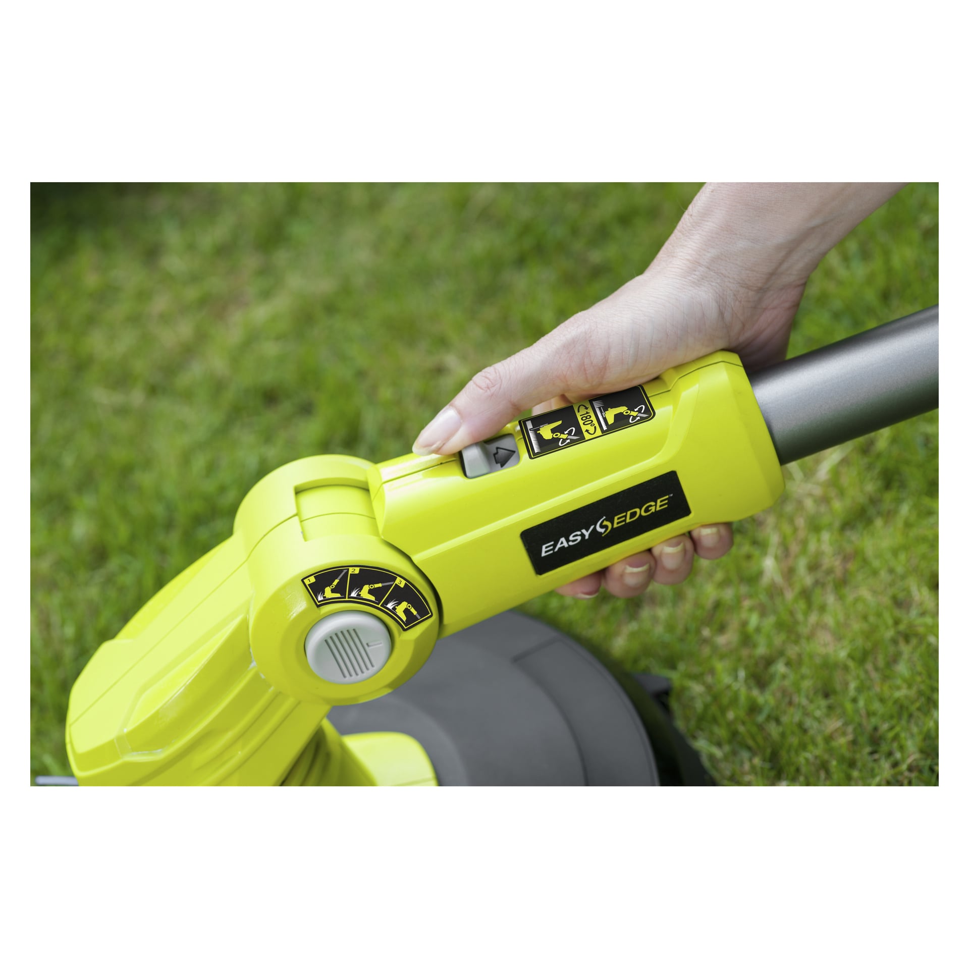 En hand justerar inställningarna på en grön Ryobi One+ OLT1832A batteridriven grästrimmer medan man står på gräset. Enheten har instruktionsetiketter som säger läsmanualen och ögonskydd krävs, med en svart knapp bredvid handtaget.