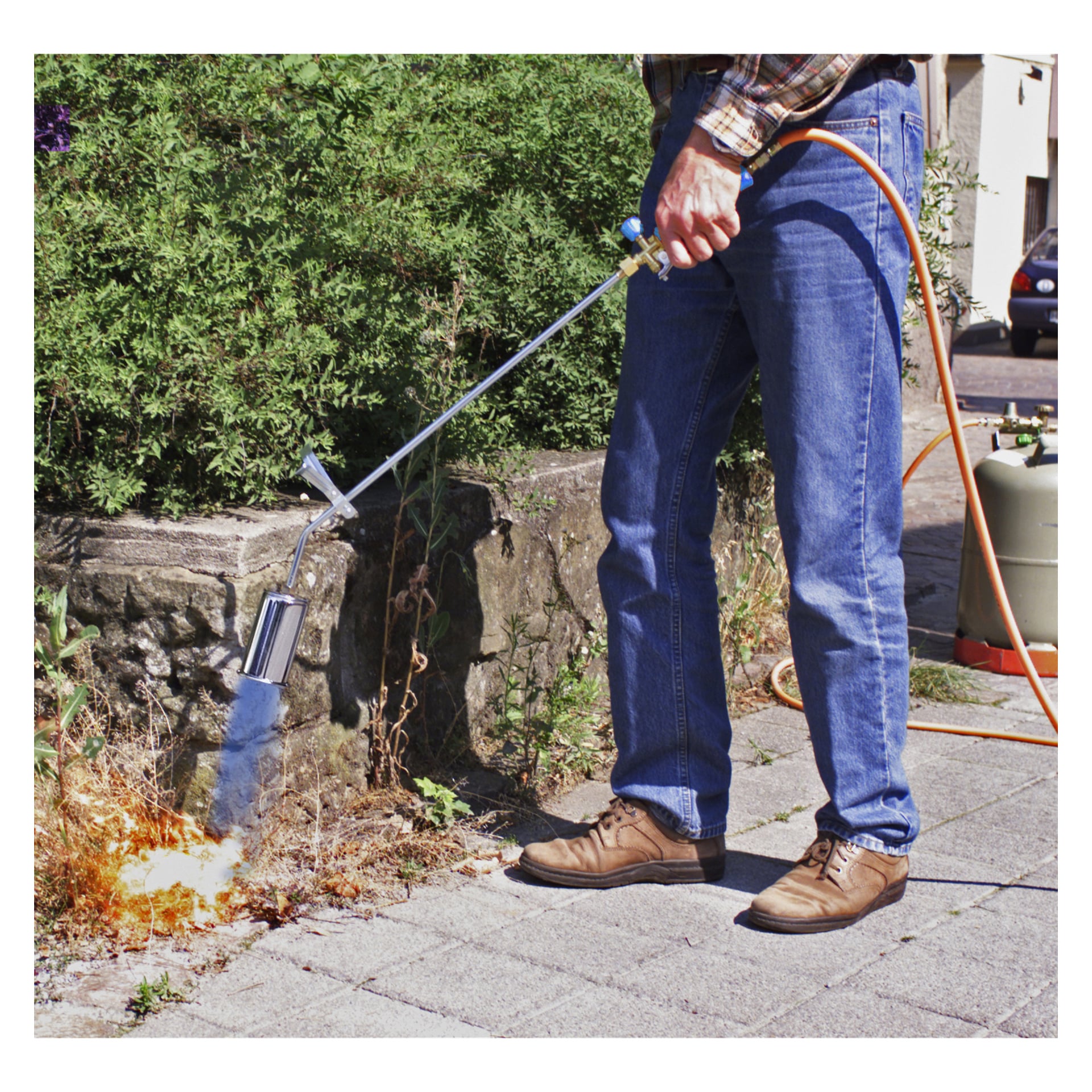 I jeans och bruna skor manövrerar man CFH LPG blowpipe ST 1000 som är kopplat till en propantank med en orange slang, och brinner ogräs längs en trottoar med gröna buskar och en stenvägg i bakgrunden.