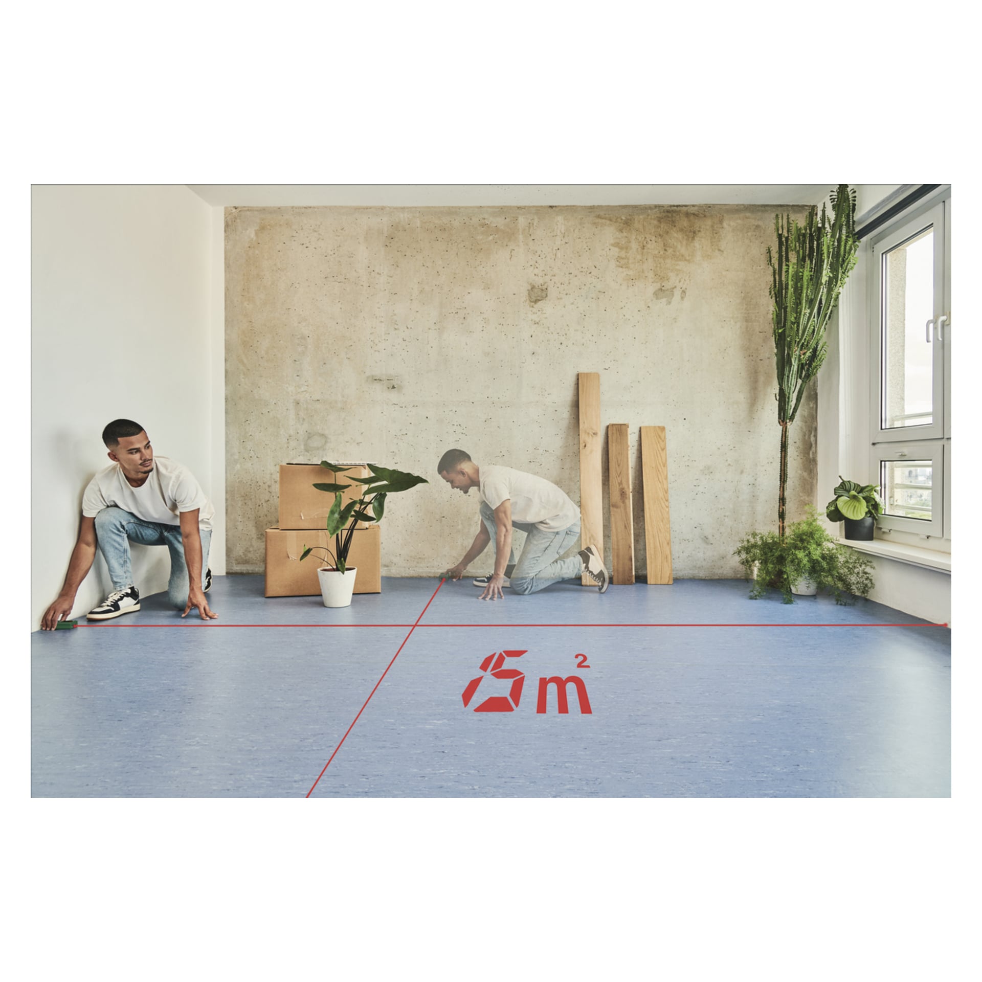 Mężczyzna mierzy powierzchnię 6 m² za pomocą miernika odległości Bosch EasyDistance 20 w pokoju z niebieską podłogą, kartonowymi pudłami, deskami i kilkoma zielonymi roślinami przy oknie przy betonowej ścianie.